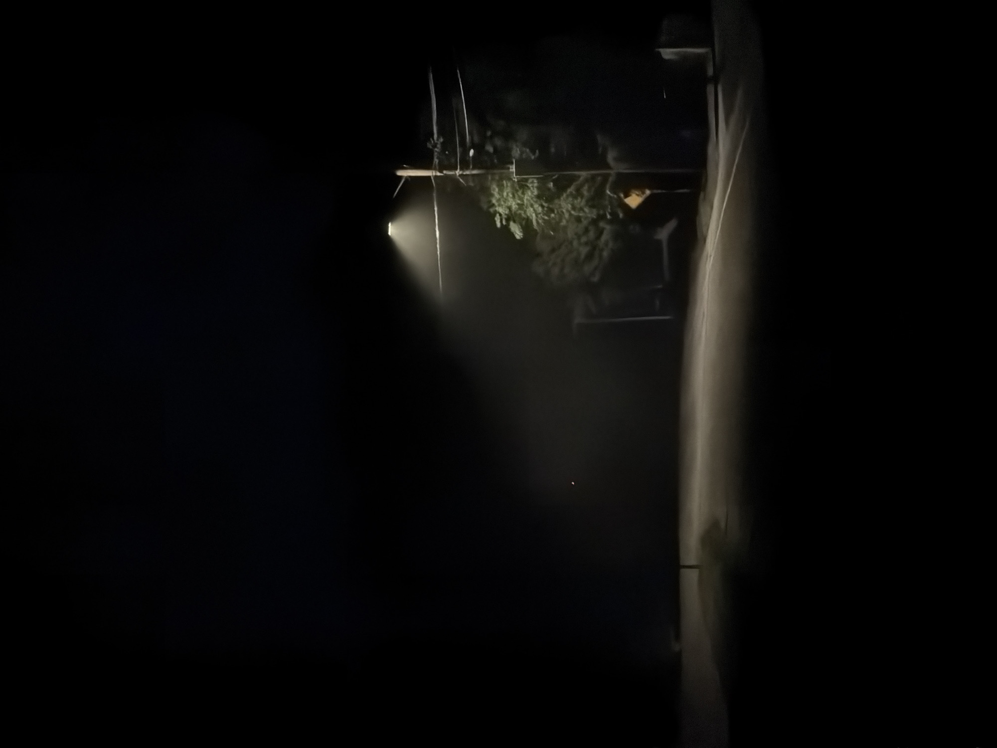 A nighttime photograph depicts a dimly lit, rural road in Hampton Bays, New York. A single streetlamp illuminates a small section of the asphalt, casting a hazy glow through what appears to be fog or mist. The road curves gently to the right, disappearing into the darkness. To the right of the road, a wooden utility pole stands, with wires strung across it and a streetlamp attached. Adjacent to the pole, a cluster of trees and foliage are visible, their shapes obscured by the low light. A yellow diamond-shaped sign is mounted on a post near the trees. The sky above is completely black, indicating the absence of natural light. No people or vehicles are present in the scene. The overall impression is one of quiet desolation.