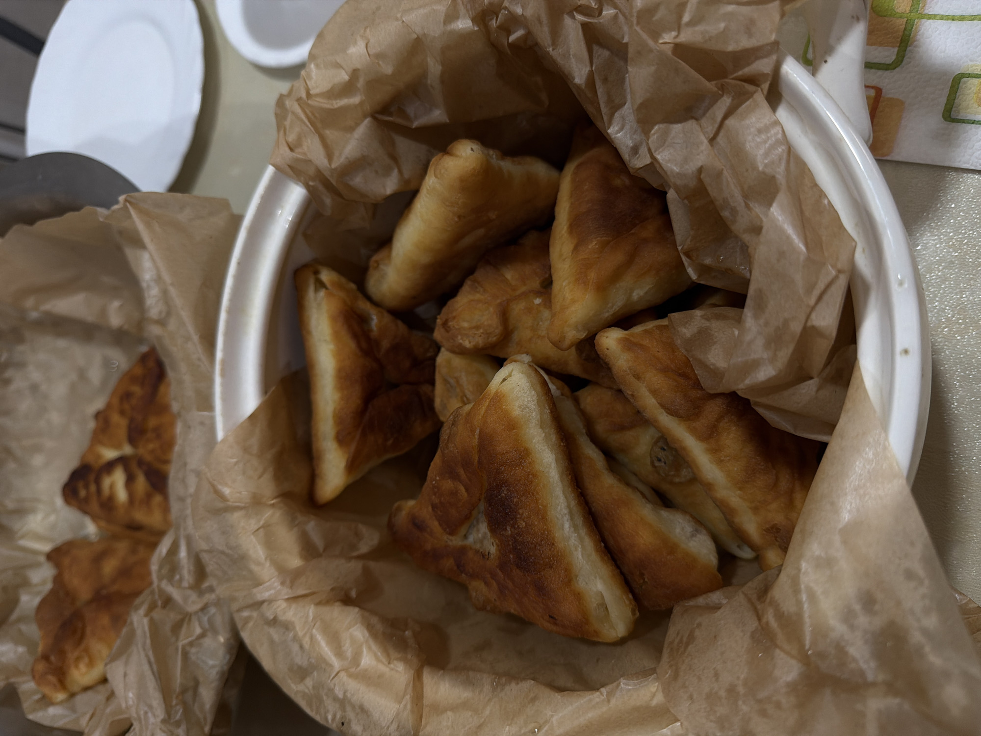 The image depicts a white bowl filled with triangular fried pastries, possibly pirozhki or samsa, lined with brown paper. Another batch of pastries is in a container to the left. Two white plates are partially visible in the upper left corner. The background appears to be a kitchen countertop. The location is specified as Chaikovsky, Russia.