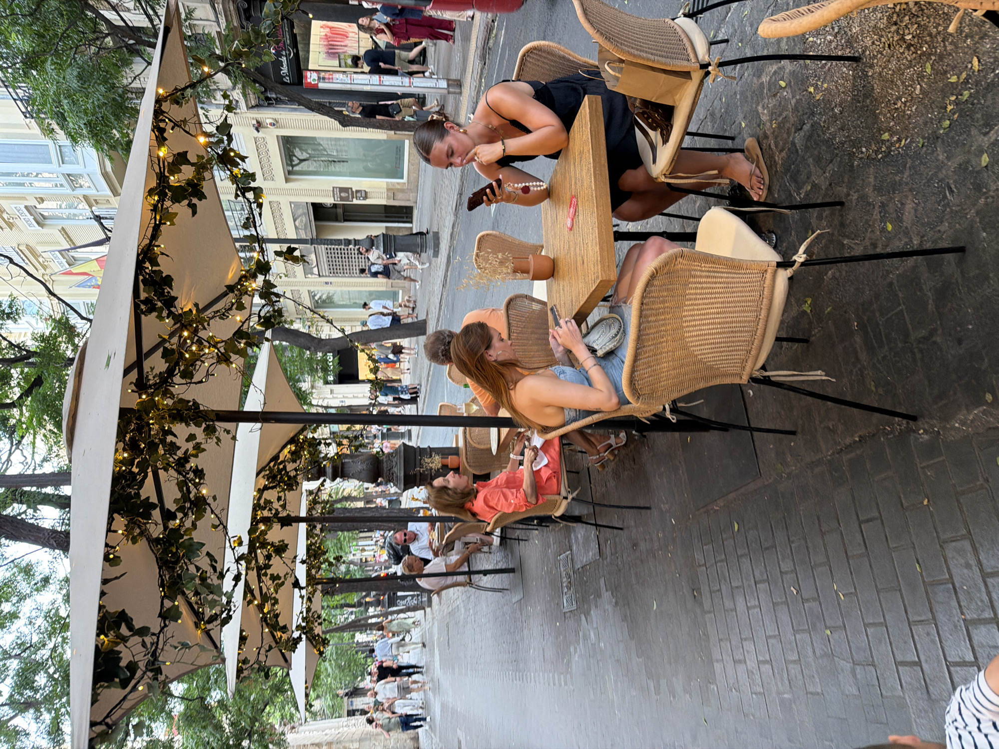 The image depicts an outdoor seating area of a cafe or restaurant on a city street. Several patrons are seated at tables beneath large umbrellas adorned with string lights and ivy. In the foreground, two young women are seated at a wooden table. One is looking at her phone, holding it in her left hand, and has a necklace with pearls and a charm. The other woman is looking down at her phone, which is on the table. A third woman in an orange dress is seated at a table further back, facing left, holding a paper in her hand. In the background, pedestrians are walking along the sidewalk, and buildings with balconies line the street. The ground is paved with cobblestones.