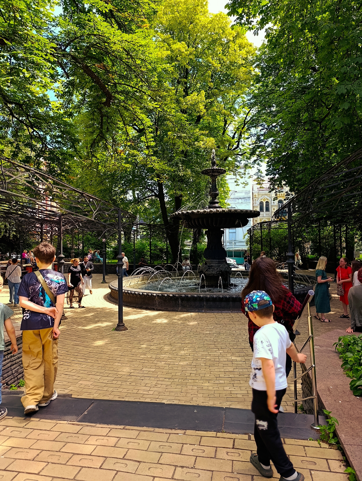 The image depicts a sunny day in a park in Kyiv, Ukraine, featuring a large, ornate fountain at its center. Lush green trees with dense foliage provide ample shade. Several people are present in the park, walking and congregating near the fountain. In the foreground, a young boy wearing a white t-shirt, black pants, and a colorful cap walks down a paved pathway. To his left, another boy in a dark t-shirt and tan pants stands facing away from the camera. Behind them, a woman in a red dress and a woman in a green dress are engaged in conversation near the fountain. The park features wrought-iron pergolas and a brick-paved walkway. In the background, buildings with architectural details are visible beyond the trees.