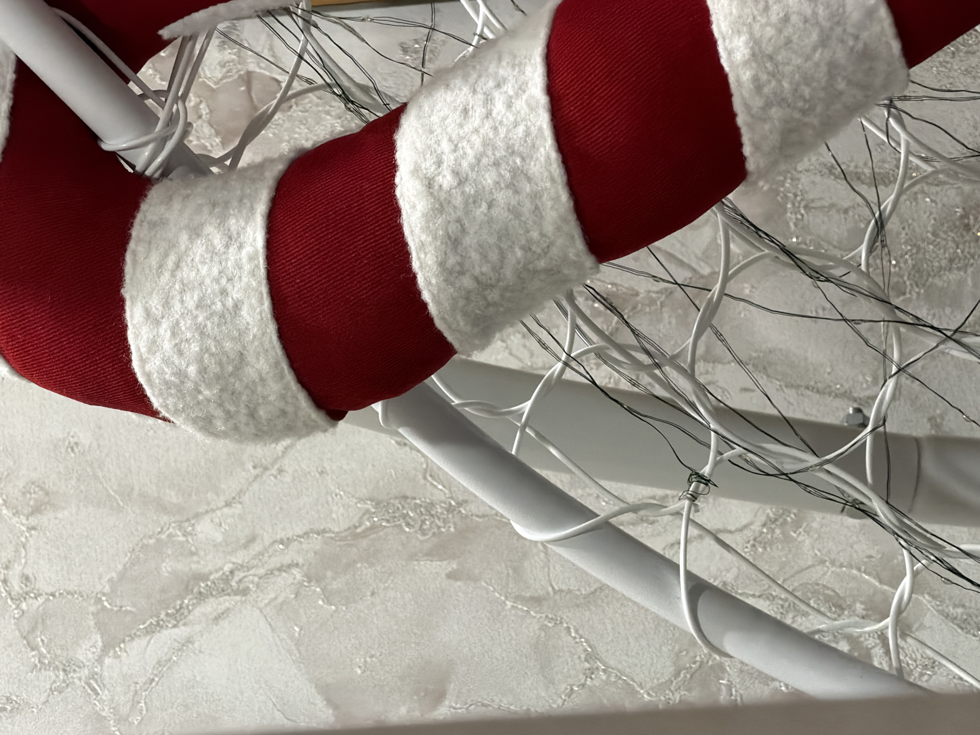 The image displays a close-up view of a large, festive decoration. A prominent, curved object, resembling a candy cane, occupies the right side of the frame. This object is wrapped in solid red fabric, interspersed with thick, white, plush bands, approximately 10-15 cm wide, creating a striped pattern. It appears to be made of a soft material.
Behind and partially intertwined with this candy cane-like object is a white, tubular or multi-wired structure, forming a decorative frame. Numerous thin, dark-colored wires, indicative of string lights, are haphazardly wrapped around this white framework. Some white wires are also visible, possibly integrated into the frame or additional lighting.
The background consists of a light-toned wall with a textured, possibly stucco or marble-effect finish, exhibiting irregular, slightly raised patterns with varying reflectivity. No people or specific actions are visible. The scene suggests an indoor setting. The precise location within the city of Perm, Russia, cannot be determined from the visual information provided in the image.