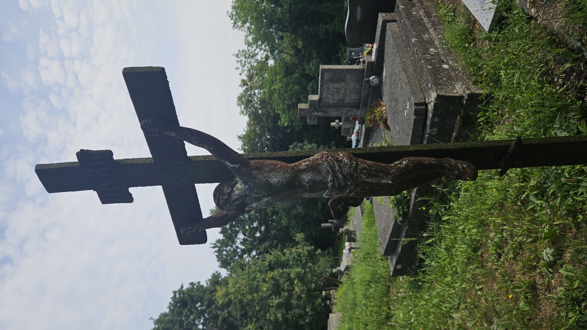 The image depicts a close-up view of a weathered crucifix in a cemetery, set against a cloudy sky and a backdrop of lush green trees. The crucifix, made of a dark, possibly stone or metal material, is adorned with a figure of Jesus Christ in a state of deterioration, showing signs of age and exposure to the elements. The figure of Jesus is depicted with a loincloth and arms outstretched.
In the background, several gravestones and mausoleums are visible, arranged in rows within the cemetery. The ground is covered in grass and some scattered wildflowers. The overall atmosphere is serene and somber, suggesting a place of remembrance and rest. This cemetery is likely located in the city of Prešov, Slovakia, given the subject matter and context.