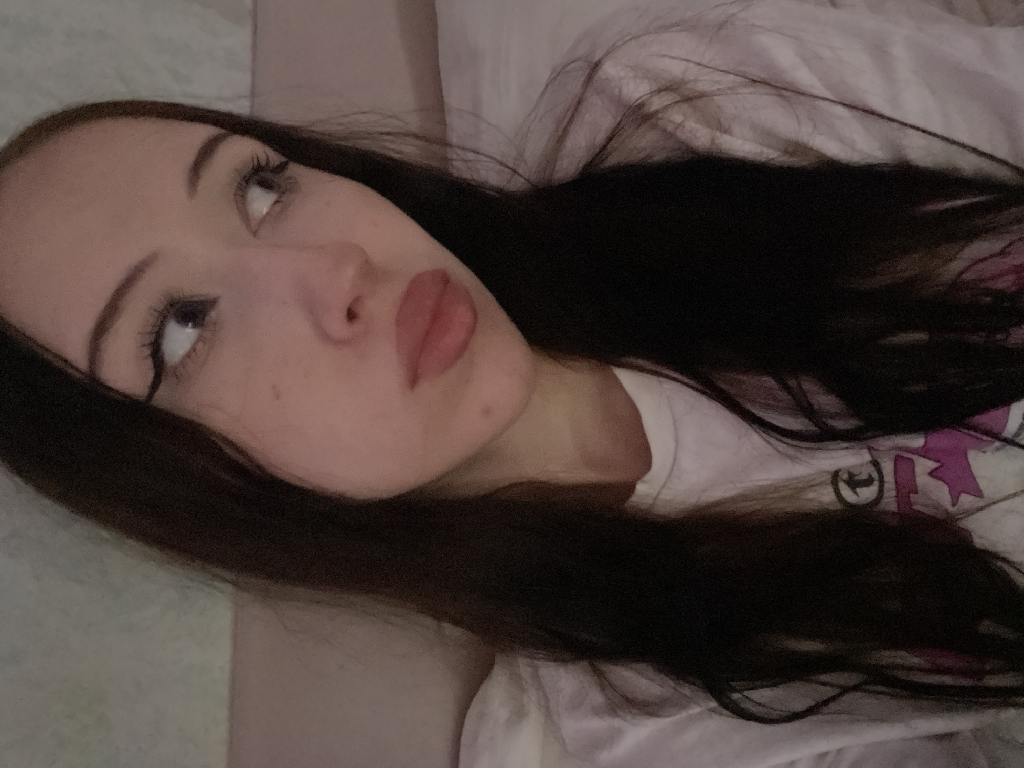 This is a close-up, top-down, over-the-shoulder shot of a young woman lying down, looking upwards and to her left. Her dark brown hair is spread out around her. She is wearing a white t-shirt with a graphic design featuring a pink shape and some black lettering, possibly a logo. The woman has fair skin, full lips with a reddish tint, dark eyebrows, and long, dark eyelashes. Her eyes are wide open and looking upwards. The scene appears to be indoors, possibly a bedroom, with a soft, possibly white, textured surface beneath her. The lighting is dim, suggesting it could be nighttime or a room with low light. The overall mood is calm and introspective. No specific text is clearly legible beyond what is on her t-shirt.