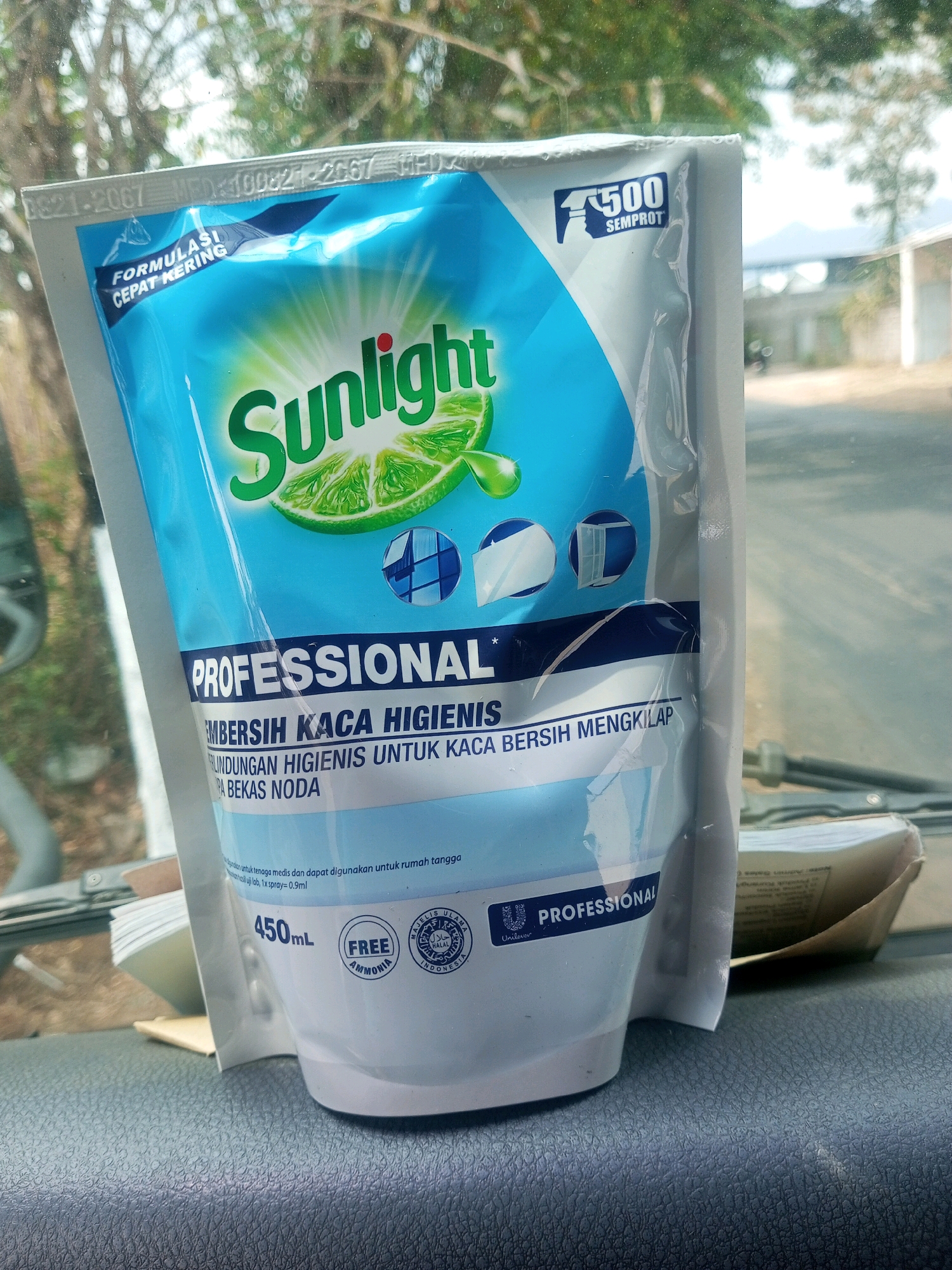 A close-up, eye-level shot shows a blue and white pouch of Sunlight Professional glass cleaner. The pouch is partially obscured by a clear plastic film, and it is placed on a textured dark gray surface that appears to be the dashboard of a vehicle. In the background, through a window, a blurred outdoor scene is visible, with trees and buildings under a bright sky, suggesting a location in Pakis, Indonesia. The Sunlight pouch features branding with a lime graphic and text in Indonesian, including "FORMULASI CEPAT KERING" (Fast Drying Formula), "PROFESIONAL," and "PEMBERSIH KACA HIGIENIS" (Hygienic Glass Cleaner). A small seal with "FREE AMMONIA" and a halal certification mark is also visible. The pouch's volume is indicated as "450mL."