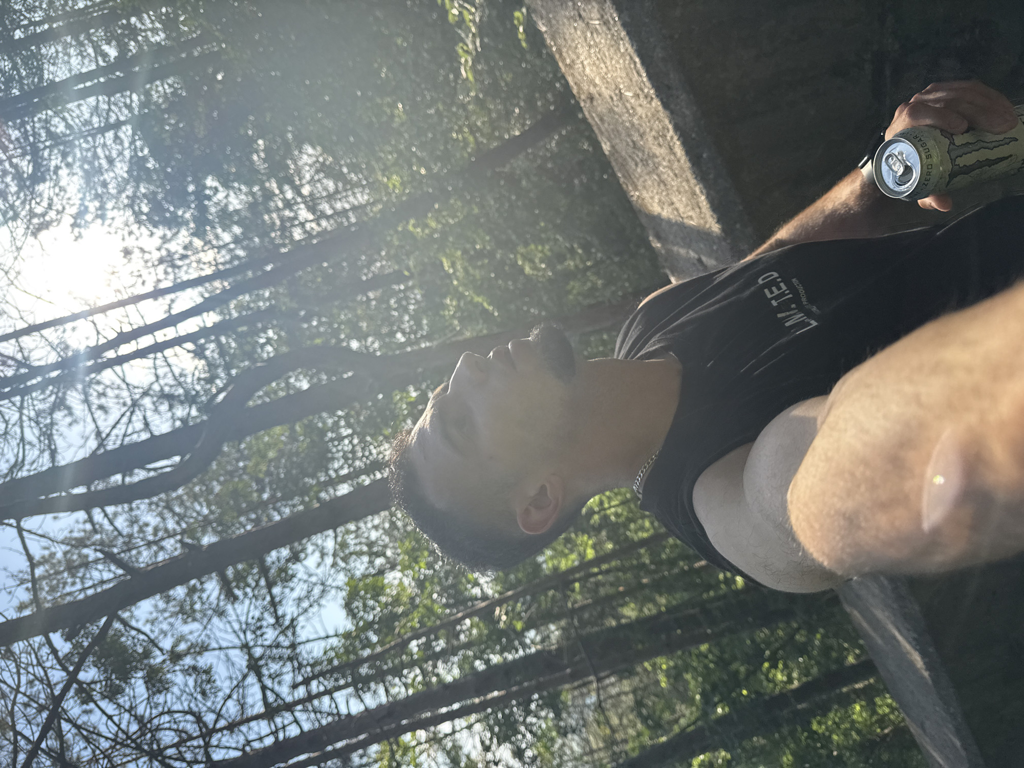 A man stands outdoors in a wooded area in Shumen, Bulgaria, holding a can of Monster Energy drink. He is positioned near a concrete structure, possibly a bridge or wall. The man is wearing a black t-shirt and a chain necklace. The trees above create a dappled light effect.