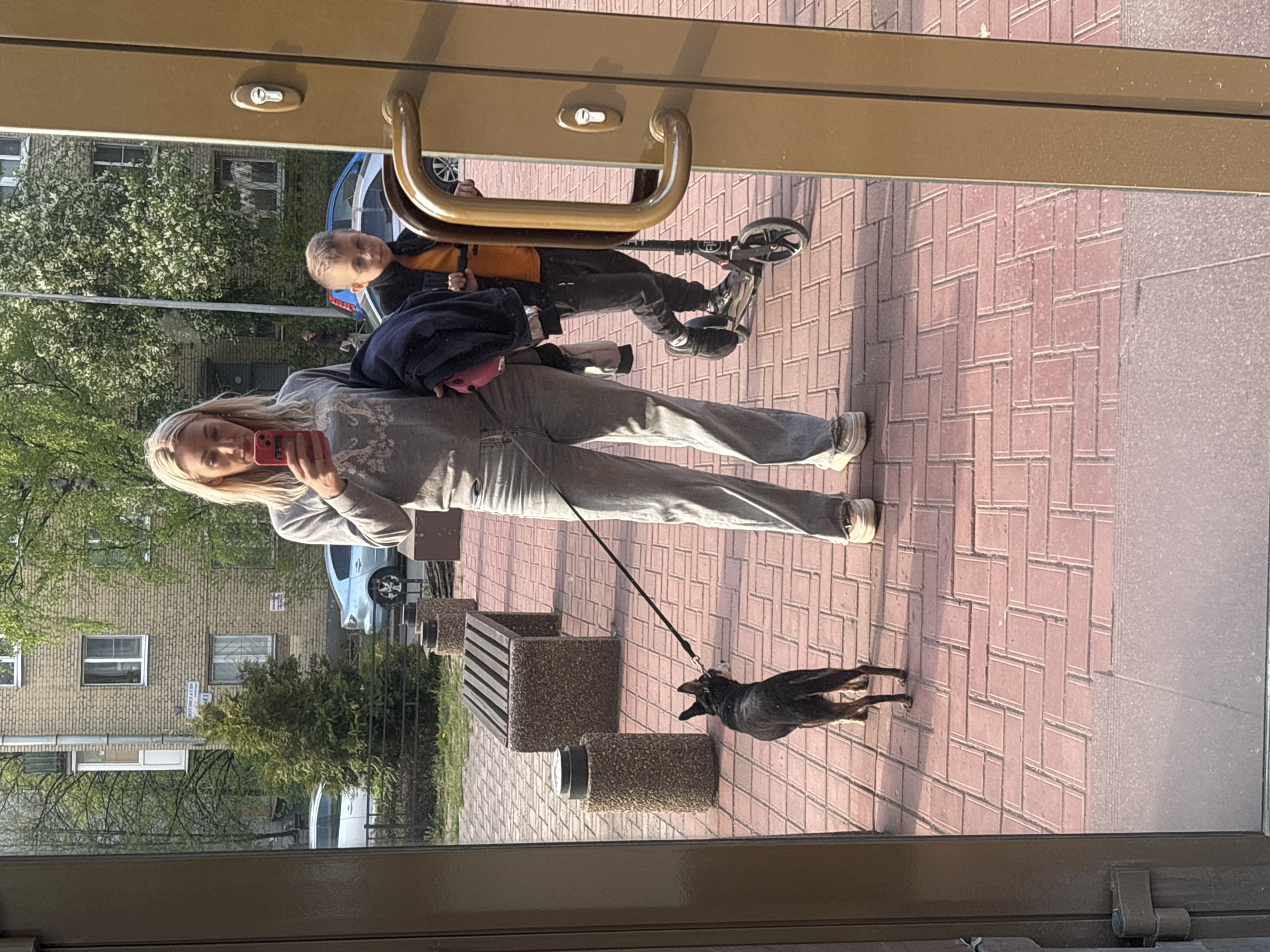 The image captures a reflection on a glass door or window, presenting an outdoor scene. A woman in a gray tracksuit holds a red phone, likely taking a photo. A young boy stands behind her on a scooter. A black dog is on a leash in front of the woman. Background features a building with light brick facade and windows, suggesting a residential area. Street furniture, including benches, are visible. The location is confirmed as Saint Petersburg, Russia, based on the street sign visible on the building.