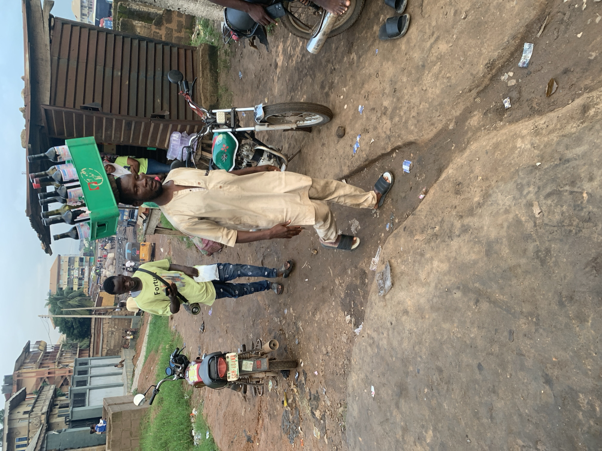 The image depicts a street scene in Shagamu, Nigeria. In the foreground, a man wearing a light-colored traditional tunic and pants, with black sandals, is walking away from the camera. He is carrying a green plastic crate filled with bottles of wine on his head. To his left, another man in a bright yellow t-shirt and dark jeans, also wearing sandals, is walking towards the camera while looking at his phone and holding a white plastic bag.
Two motorcycles are parked on the dirt road. The motorcycle on the left is red and black, and the one on the right is green and black. Behind the man with the wine crate is a small wooden structure, possibly a shop, with a corrugated metal roof. In the background, several multi-story buildings line the street. The ground is unpaved and dusty, with some debris scattered around. Other individuals and a tricycle are visible in the distance, suggesting a bustling market or residential area. The sky is overcast.