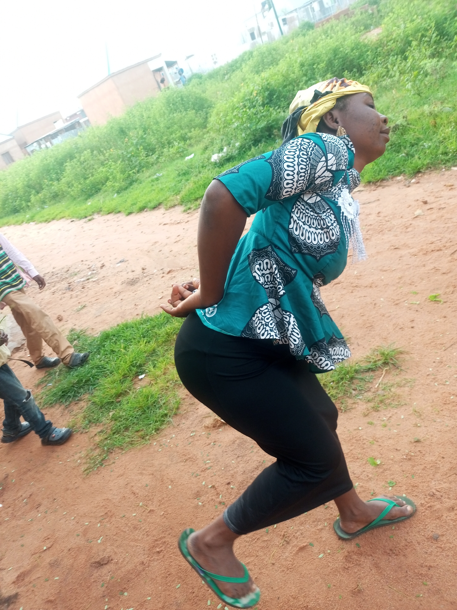 An overhead shot shows a woman in a teal and white patterned top, black leggings, and green flip-flops with her hands clasped behind her back. She is walking on a dirt path beside a grassy slope, with a few buildings and trees in the background. The setting appears to be a rural area in Tempure, Nigeria.