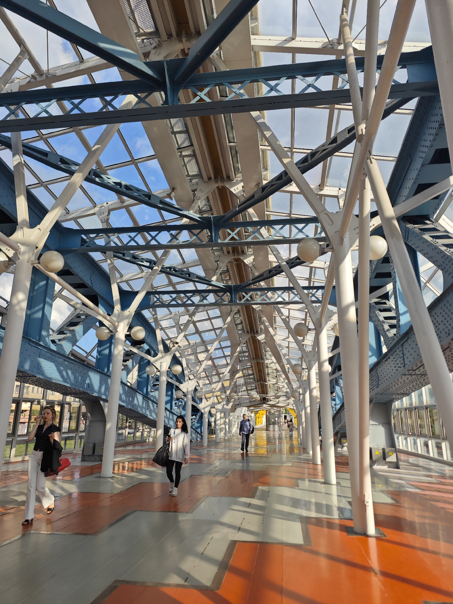The image shows the interior of a modern pedestrian bridge with a glass roof and a complex metal support structure. The bridge features white pillars that branch out like trees, supporting a network of blue and gray metal beams and crossbeams. The walkway is paved with alternating orange and gray geometric patterns. Several people are visible walking along the bridge. In the foreground, a woman in white pants and a black polka-dot top walks towards the camera, holding a red bag and a black jacket over her arm, and speaking on her phone. Further down the walkway, a woman in a white hoodie and black pants walks away from the camera, carrying a large black bag. Another man in a blue jacket walks in the distance. The architecture suggests a contemporary urban setting, likely designed for pedestrian access over a significant thoroughfare or between buildings. While the specific location is not identified in the image, the architectural style is consistent with modern public infrastructure found in large cities.