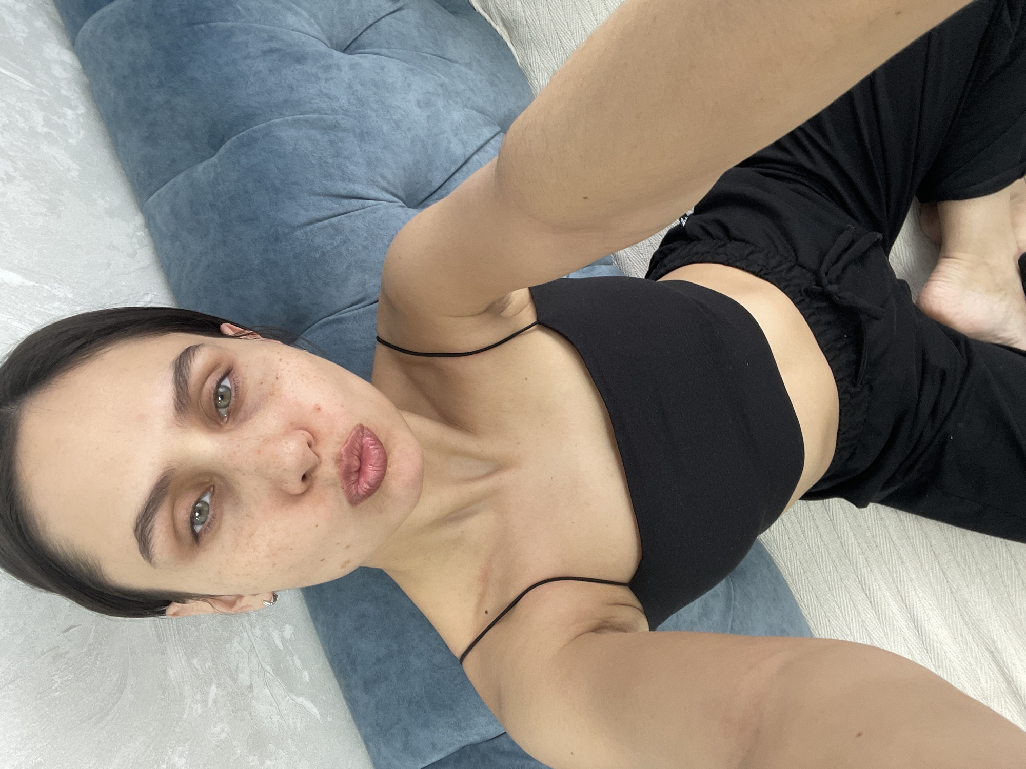 A fair-skinned woman with freckles is lying on her back on a light-colored patterned surface, possibly a bed or couch. She is wearing a black crop top with thin straps and black drawstring pants. Her dark hair is pulled back. She is looking at the camera with puckered lips. Part of a blue cushion is visible in the upper left corner of the image. The image appears to be a selfie taken with a handheld device. The location of Izhevsk, Russia, is not evident from the details in the image.