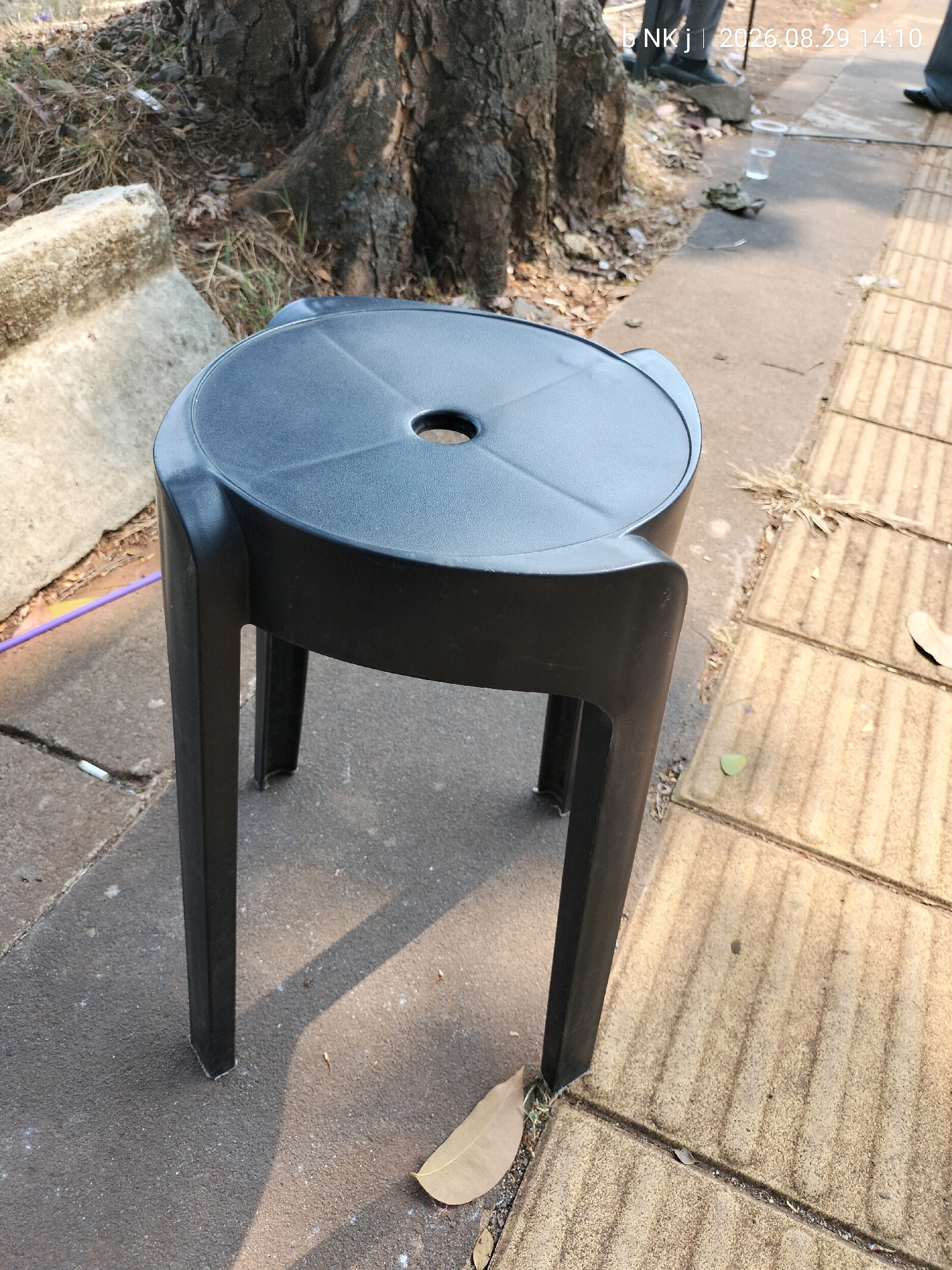 A black plastic stool stands on a paved pathway next to a large tree. The stool has four legs and a round seat with a circular opening in the center. The ground is a mix of concrete and dirt, with some leaves scattered around. In the background, people are visible walking and standing. A clear plastic cup with a small amount of liquid is on the pathway. The scene appears to be outdoors, likely in a public area. The timestamp overlay indicates "b NK j | 2026.08.29 14:10".