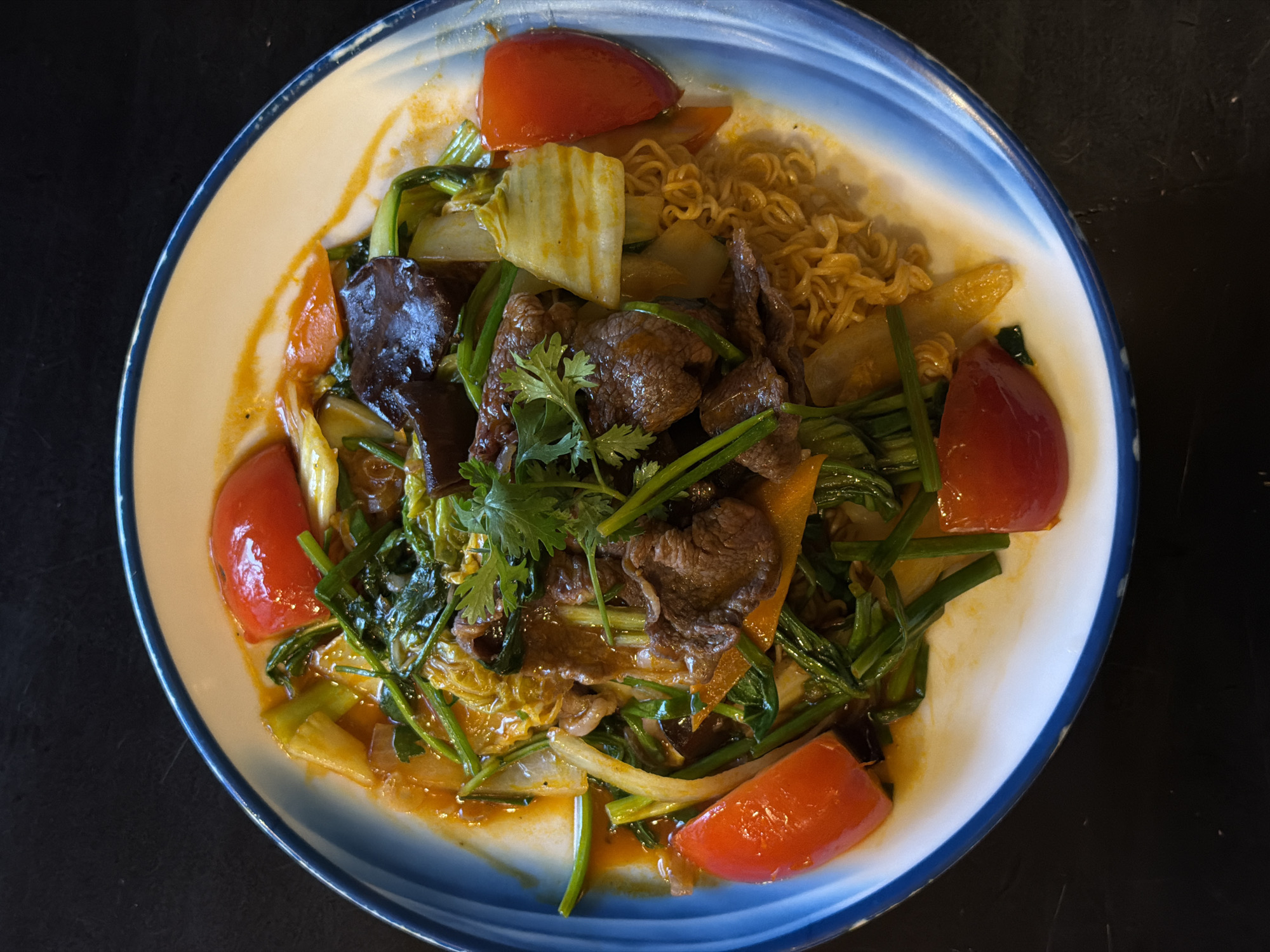 A top-down shot shows a plate of Vietnamese food served in Đà Nẵng, Vietnam. The dish, arranged on a plate with a light-colored center and a blue rim, features an arrangement of sliced tomatoes, braised beef, noodles, various green vegetables, and black fungus mushrooms, all drizzled in sauce. The overall composition suggests a traditional Vietnamese dining experience.