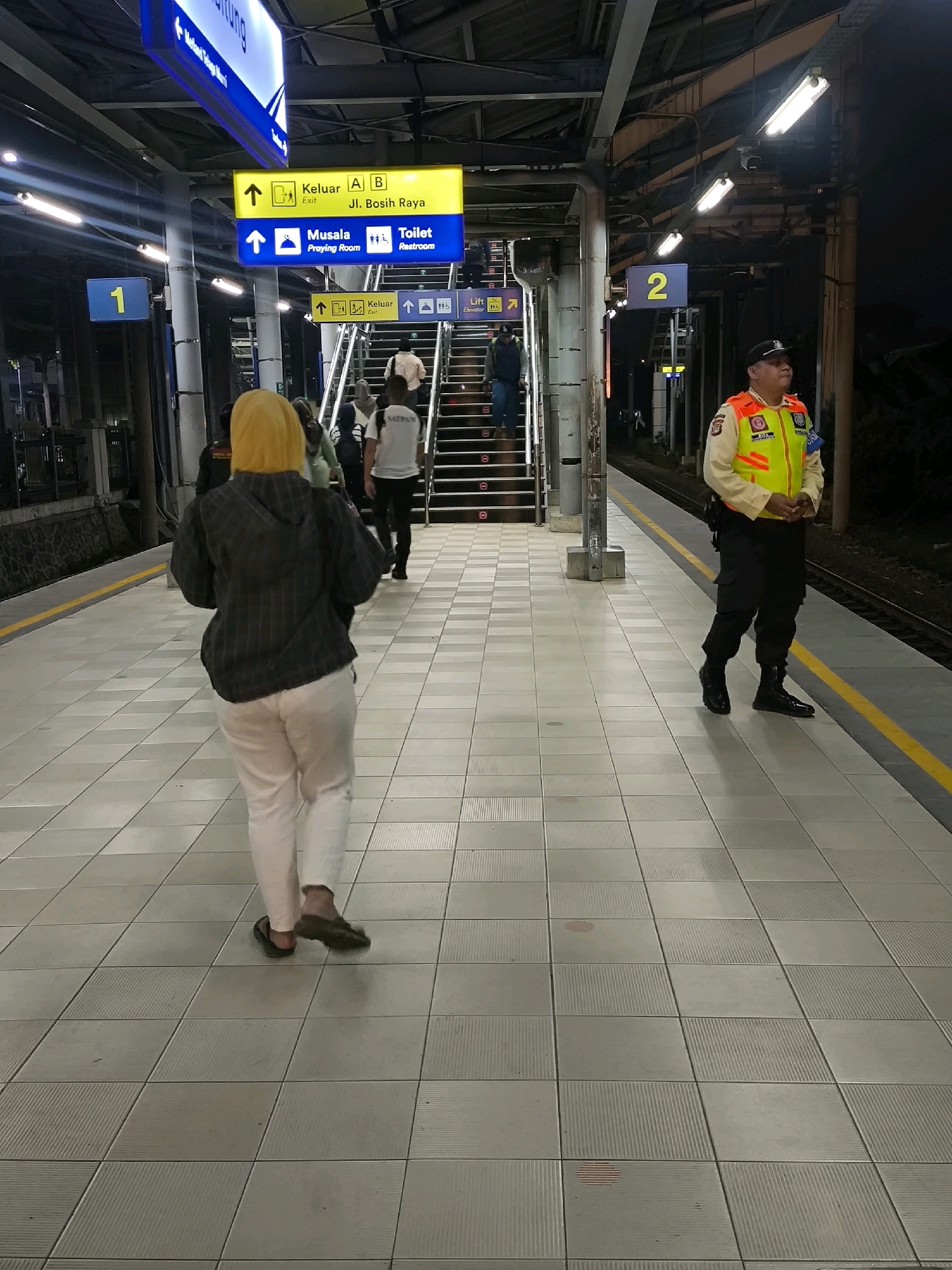 The image depicts a train station platform in Mekarsari, Indonesia. A person with bright yellow hair is walking away from the camera on a tiled platform. They are wearing a dark checkered jacket and white pants. To the right, a uniformed security guard in a reflective vest stands facing away from the camera. Yellow lines mark the edge of the platform. Overhead signs indicate directions to "Exit," "Musala (Praying Room)," and "Toilet (Restroom)," along with platform numbers 1 and 2. Stairs leading to a higher level are visible in the background, with several people ascending them. The lighting suggests it is evening or night.