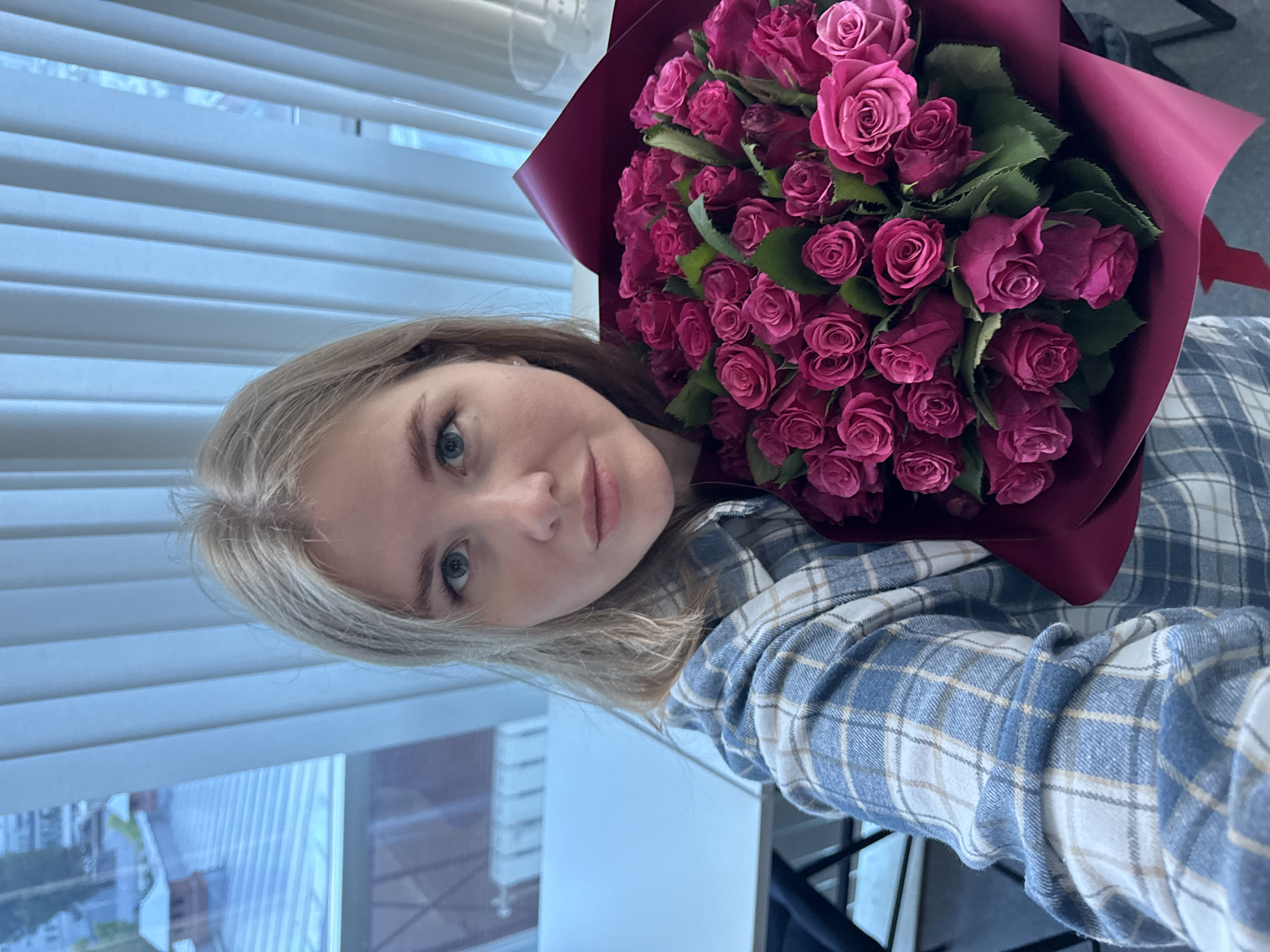 A young woman with blonde hair and blue eyes is holding a bouquet of dark pink roses. She is wearing a blue and white plaid shirt. The background appears to be an indoor setting with vertical blinds covering a window, through which the rooftops of buildings can be seen, suggesting an urban environment. The lighting is soft and diffused. There is no information in the image to suggest a specific location in the city of Perm, Russia.