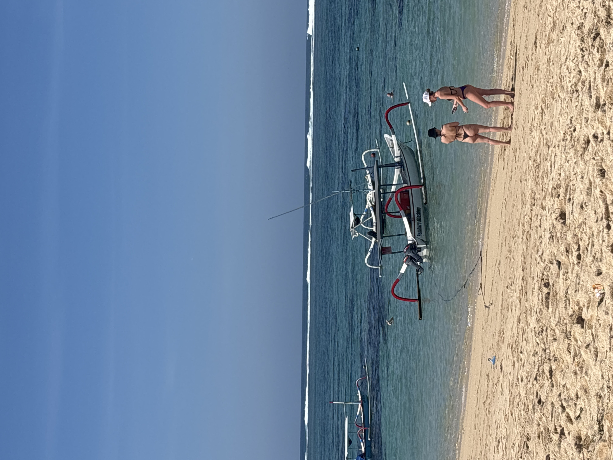 The image depicts a tranquil beach scene in Kuta Selatan, Indonesia. Two women in bikinis are standing on the sandy shore, facing the ocean. The woman on the left wears a black bikini, while the woman on the right is in a purple bikini and a white cap. Behind them, traditional Indonesian fishing boats are anchored in the clear turquoise water. One boat, prominently featured, is white with red outriggers and has an outboard motor. The ocean exhibits gentle waves breaking further out. The sky is a clear, bright blue, with no clouds visible. The sand is light brown and shows some texture from the tide. The overall atmosphere is one of a relaxed, sunny day at the beach.