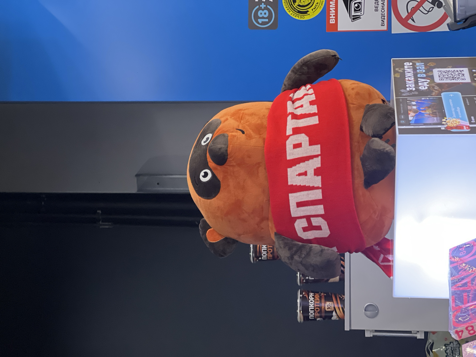 The image captures a plush toy of a raccoon-like character wearing a red scarf with the word "СПАРТАК" (Spartak) in white letters. The toy is positioned in front of a blue background and appears to be at an event or promotional display. To the left of the toy, there are several dark cylindrical containers with labels that include Russian text such as "ПОПКОРН" (popcorn) and "ПРОТЕИН" (protein). In the lower left corner, a white illuminated box with a screen displays promotional content. To the right of the toy, there are signs with Russian text, including one with an "18+" symbol and another with a circular emblem and text that reads "ОБЪЕКТ НА НАХОЖДЕНИИ" (Object in the area). The overall impression is a retail or entertainment venue, possibly a cinema or a food court, in Moscow, Russia.
