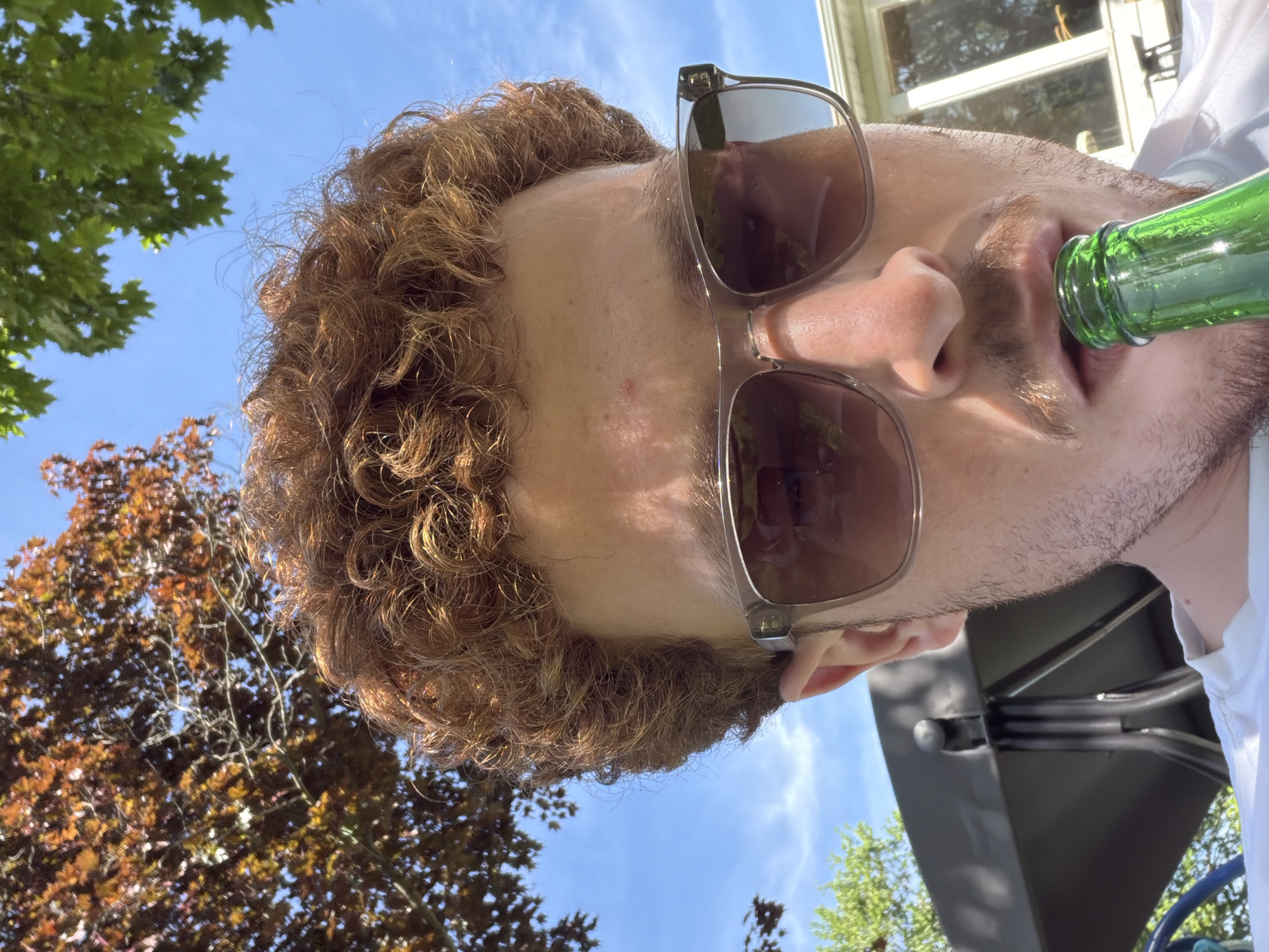 The image, taken in Pepper Pike, United States of America, captures a close-up, low-angle perspective of a person outdoors on a bright, sunny day. The subject is a male with reddish-brown curly hair and light facial stubble, wearing dark sunglasses. The image is oriented sideways, providing an upward-looking view.
The primary activity shows the person drinking from a green glass bottle, which is held to their mouth. The dark lenses of the sunglasses reflect the blue sky and surrounding foliage. The background features a clear blue sky with sparse white clouds, indicating fine weather. Lush green deciduous trees are visible on the left side of the frame, while trees with reddish-brown foliage appear towards the bottom, suggesting either late summer, early autumn, or specific tree varieties. A building structure, possibly a house with a window, is partially visible in the upper right, and a dark outdoor structure or furniture piece is in the lower right.
The scene conveys a casual and relaxed outdoor moment, likely in a backyard or patio setting. The bright sunlight creates strong highlights on the person's hair and skin, as well as on the leaves of the trees. No visible text is present in the image.