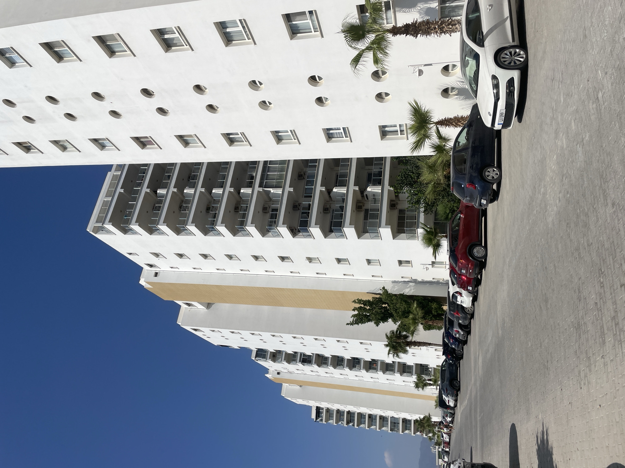 A daytime, eye-level, full shot captures a row of modern, white apartment buildings in Trikomo, Northern Cyprus. The buildings are tall with multiple stories, featuring balconies with glass railings and a central section accented with a golden-yellow facade. In front of the buildings, a paved parking lot is filled with a variety of parked cars, including a white Volkswagen Scirocco in the foreground, a dark blue hatchback, and several other cars of different colors and models. Palm trees and other greenery are interspersed between the buildings and in the parking lot. The sky is a clear, vibrant blue, with distant mountains visible on the horizon.
