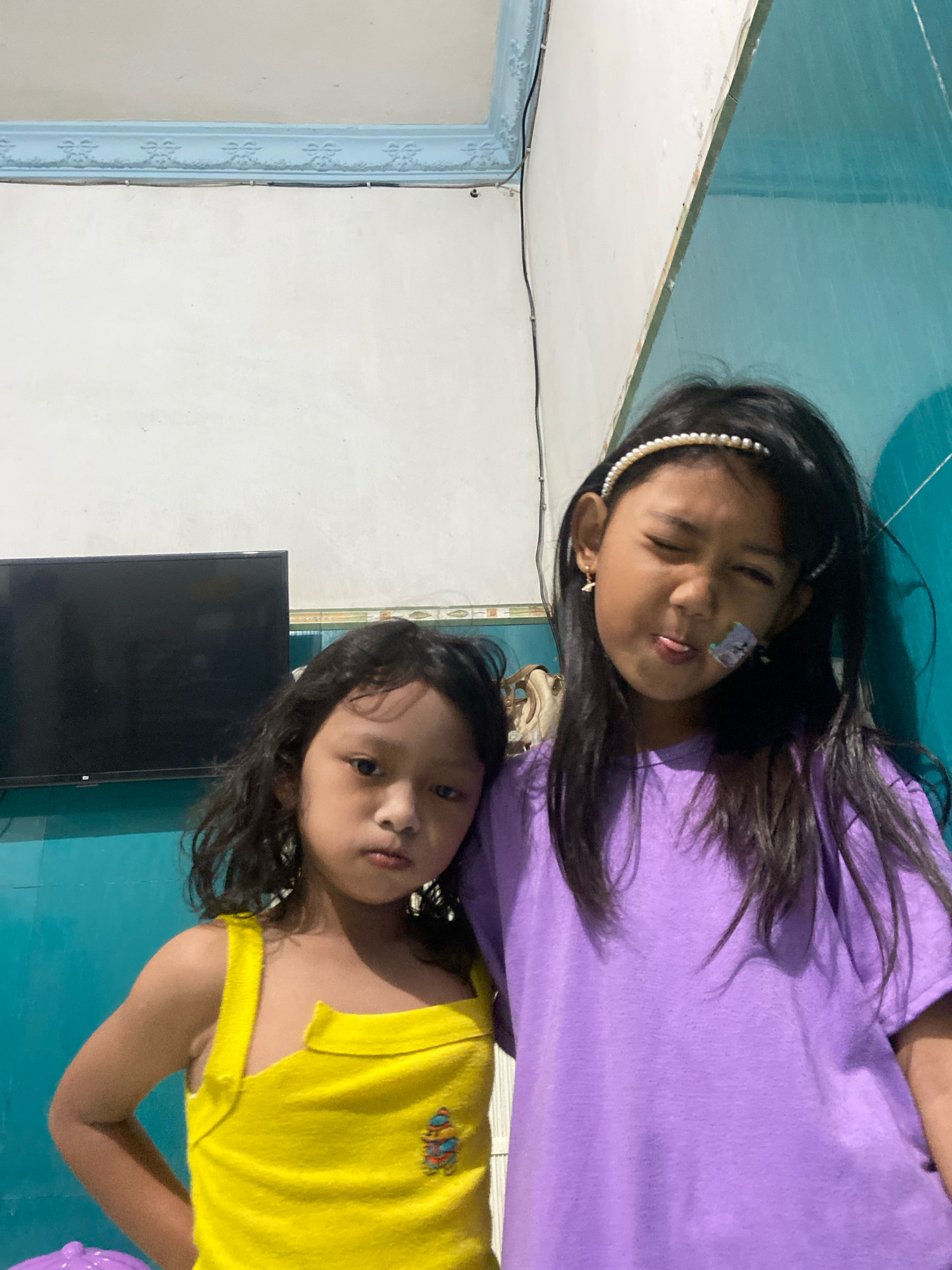 The image captures two young girls posing for a photo indoors. The girl on the left, with curly dark hair, wears a bright yellow tank top with a cartoon character embroidered on it. She has a serious expression. The girl on the right, with long dark hair and a pearl headband, is sticking out her tongue, which has a bandage on it, and winking. She is wearing a purple t-shirt and has a small gold earring in her left ear. The background shows a white wall with a decorative blue trim along the top, and a teal-colored wall on the right. A black flat-screen television is visible on the left side of the frame, mounted on the white wall. Electrical wires are present along the wall. The overall impression is a casual, home environment.