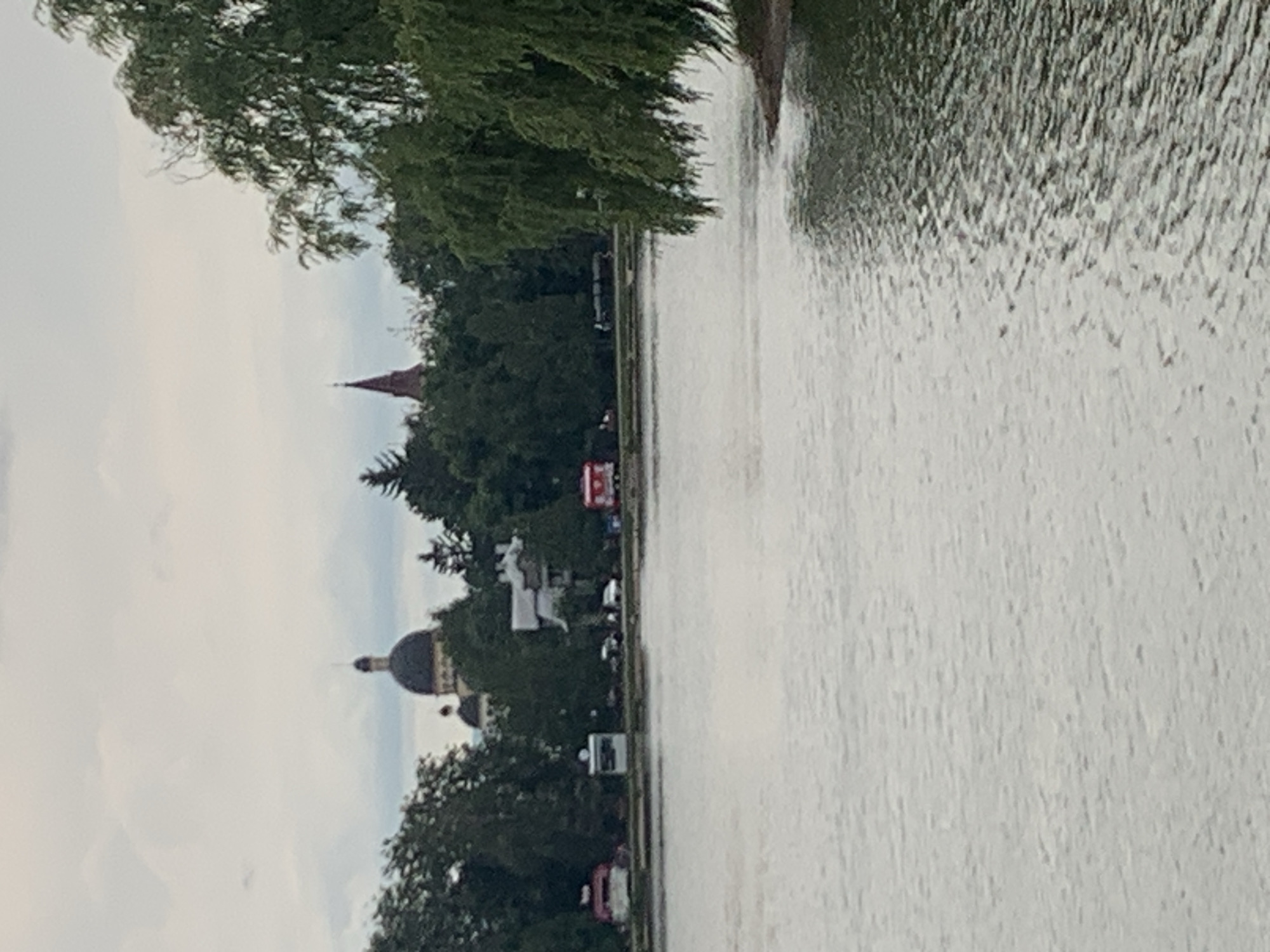 This is a medium-shot, slightly blurry, outdoor photograph taken from across a body of water, likely the Rashkiv Reservoir in Kolomyia, Ukraine. The scene is dominated by the water in the foreground, with a shoreline visible in the middle ground. Along the shoreline, there are various structures and vehicles, including what appears to be a domed building with a spire, a red-roofed building, and what looks like a red food truck or trailer. Further back, there are trees and a tall, red-pointed spire of a building. The sky is overcast with gray clouds. The water is choppy, with ripples indicating a slight breeze. The overall impression is of a park or recreational area along a body of water, with some urban or historical architecture visible in the distance.