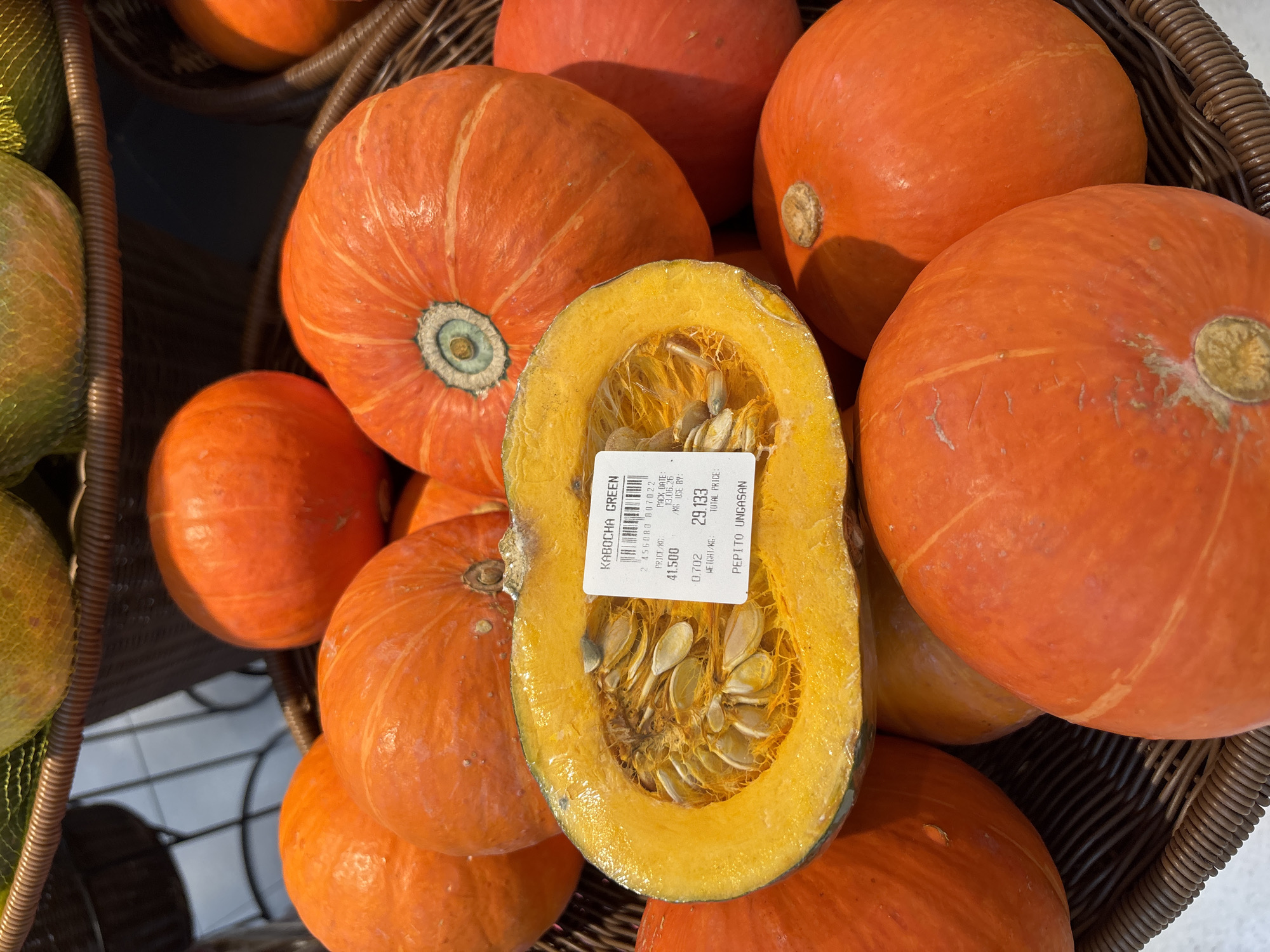 A close-up, eye-level shot shows a variety of orange kabocha squashes in a woven basket at a market. In the center of the frame, one squash is cut in half, revealing its bright yellow-orange flesh and numerous seeds. A white price sticker is attached to the cut squash, displaying the text "KABOCHA GREEN", a product code, price per kilogram, weight, and total price. The squashes are round to slightly flattened in shape, with ribbed surfaces. Some have faint greenish-yellowish stripes. The lighting is bright, highlighting the vibrant colors of the squashes. The background is blurred but suggests a market environment with hints of shelving or other produce. This image was captured in Kuta Selatan, Indonesia.