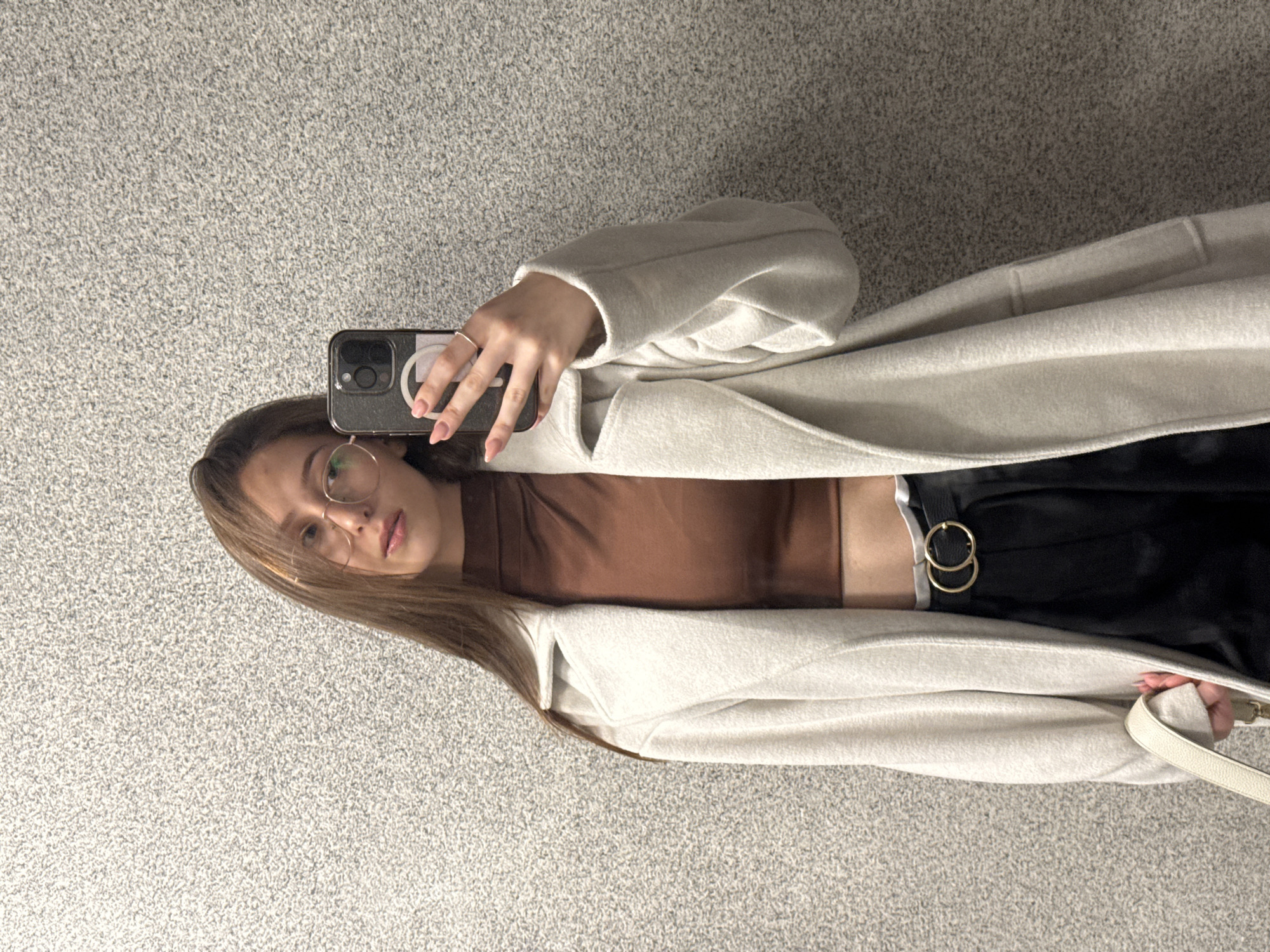 A young woman is captured in what appears to be a mirror selfie, with the image rotated 90 degrees clockwise. She has long, straight, medium-brown hair and is wearing round, thin-rimmed glasses. Her gaze is directed towards the camera with a neutral or slightly serious expression. Her lips are a light pink or nude shade, and her eyes appear to be green or hazel.
She is dressed in a light-colored, possibly off-white or cream, long coat or cardigan. Beneath it, she wears a fitted brown or terracotta-colored top and black trousers. A stylish detail on her trousers is a belt featuring two gold-colored, interlocking rings.
In her left hand, she holds a dark-colored smartphone, likely reflecting its camera lens, indicating a selfie is being taken. The phone case has a pop socket or ring holder with a partial white logo or symbol visible. Her fingernails are painted with a light pink polish, and she wears a simple silver ring on one finger. A strap from a white or cream-colored handbag is visible near her hip.
The background consists of a light gray, textured surface, possibly a carpeted floor or wall, suggesting an indoor setting. This image is likely taken in Sofia, Bulgaria.