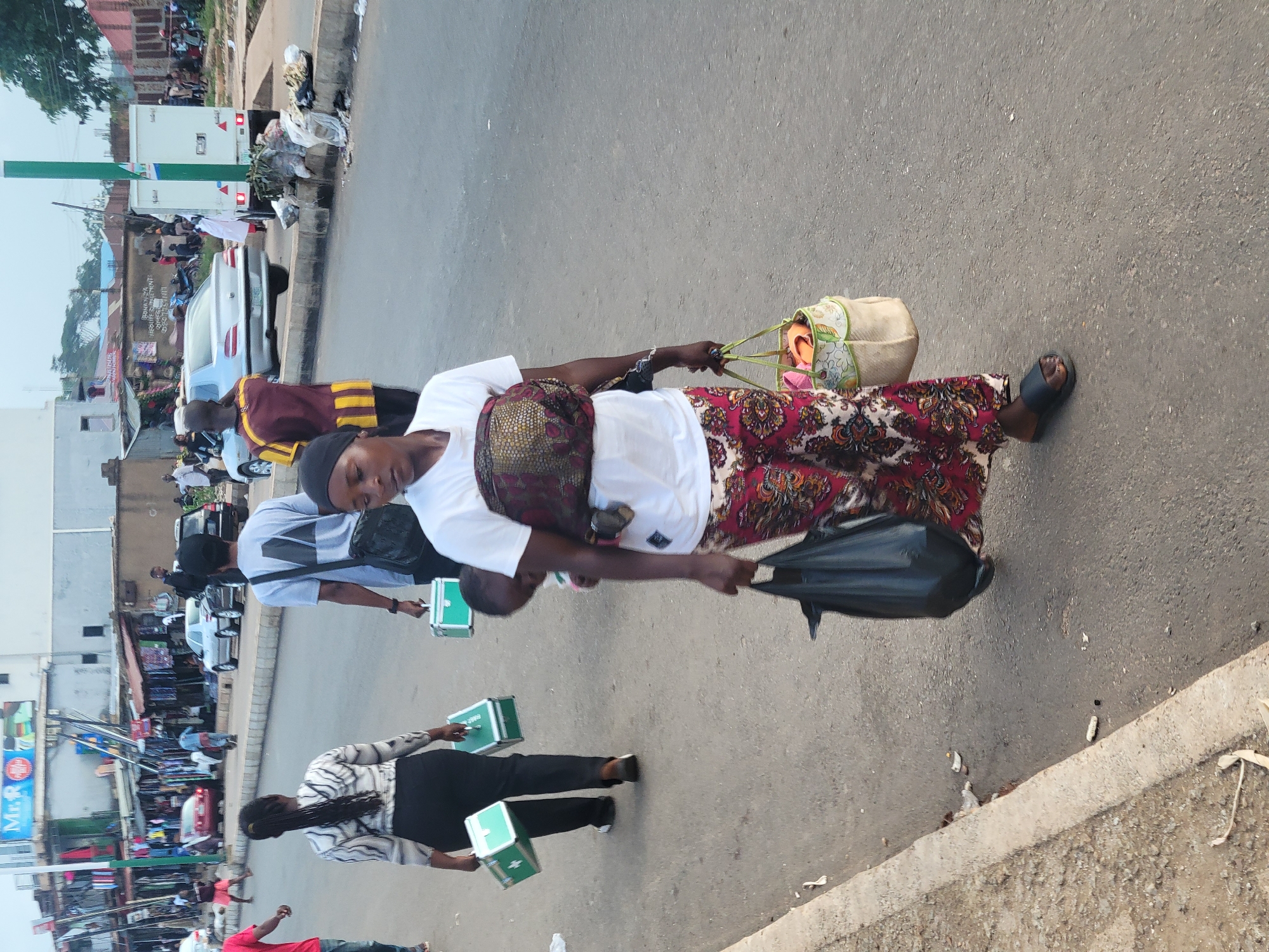 This image captures a street scene in Jos, Nigeria, likely during daylight hours. The main subjects are several people walking on a paved road, some of whom appear to be vendors or commuters.
In the foreground, a woman wearing a white shirt and a patterned skirt is carrying two bags. One is a light-colored woven bag with a decorative pattern, and the other is a large black plastic bag. She is also wearing a dark headscarf and dark sandals. Behind her, a person in a light grey shirt is seen carrying two green boxes. Further back, another individual in a black and white striped top and black pants also carries two similar green boxes.
The setting appears to be a busy urban environment with buildings and shops visible on the left side of the road. There are several vehicles on the road, including cars and a white truck. Some of the shops have displays of goods. Debris and trash are present along the edge of the road, suggesting a typical roadside environment.
The activity taking place seems to be daily commerce and movement within the city. The expressions on the faces of the people are not clearly discernible, but the overall impression is one of everyday activity. There are no overt indicators of specific weather conditions, but the lighting suggests it is daytime. The presence of the green boxes might indicate the transportation of goods or products.
A sign with the text "Mr. P" is visible on the left side of the image, possibly advertising a business.