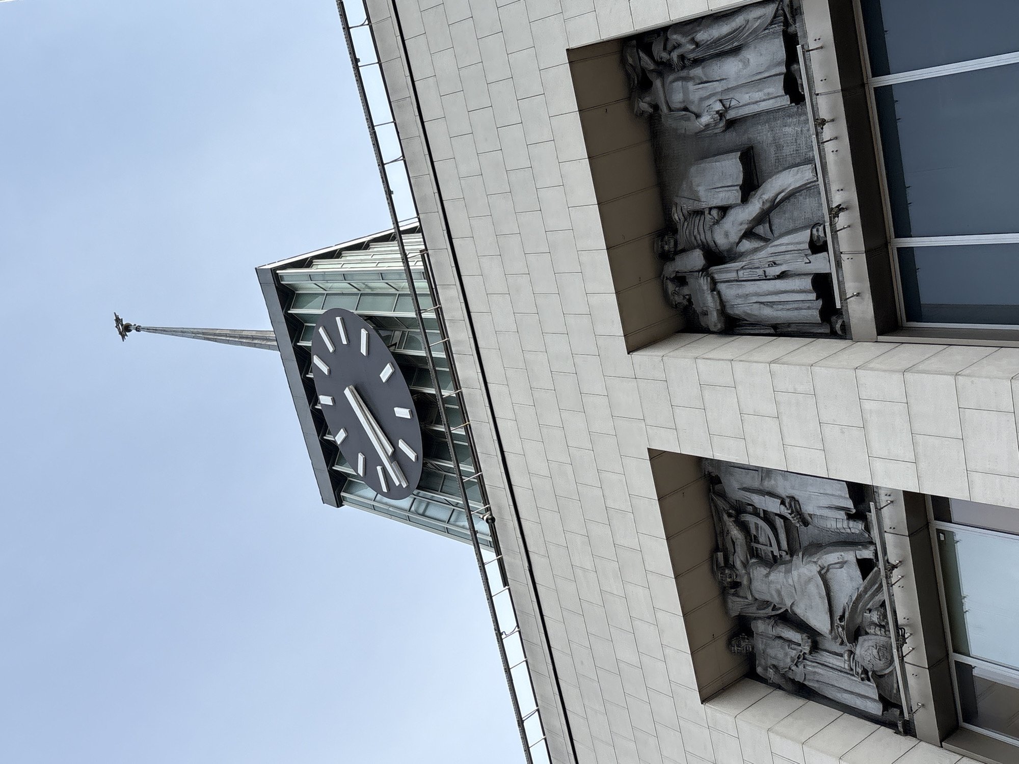 The image shows the Stockmann department store in Saint Petersburg, Russia, viewed from below. The building features a clock tower with a large black clock face. There are bas-relief sculptures on the building's facade. The sky is visible in the background.