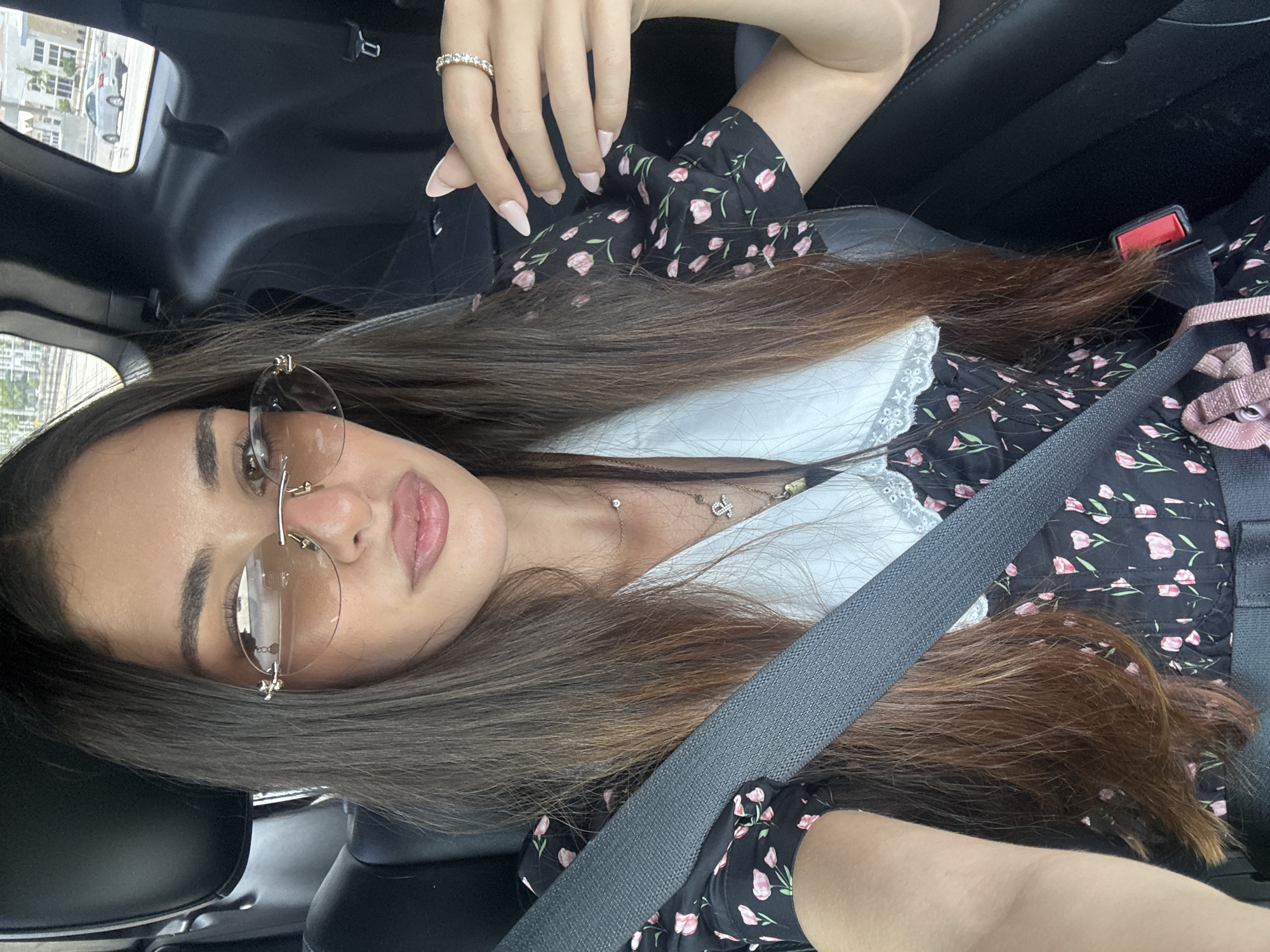 A woman with long, brown hair, wearing a black floral dress with a white collar and sunglasses, is seated in the passenger seat of a car. She is wearing a seatbelt. Her left hand is resting on her lap, while her right hand is raised, showing a ring with diamonds on her finger. The background shows the interior of the car and a blurred view of a city street with cars.