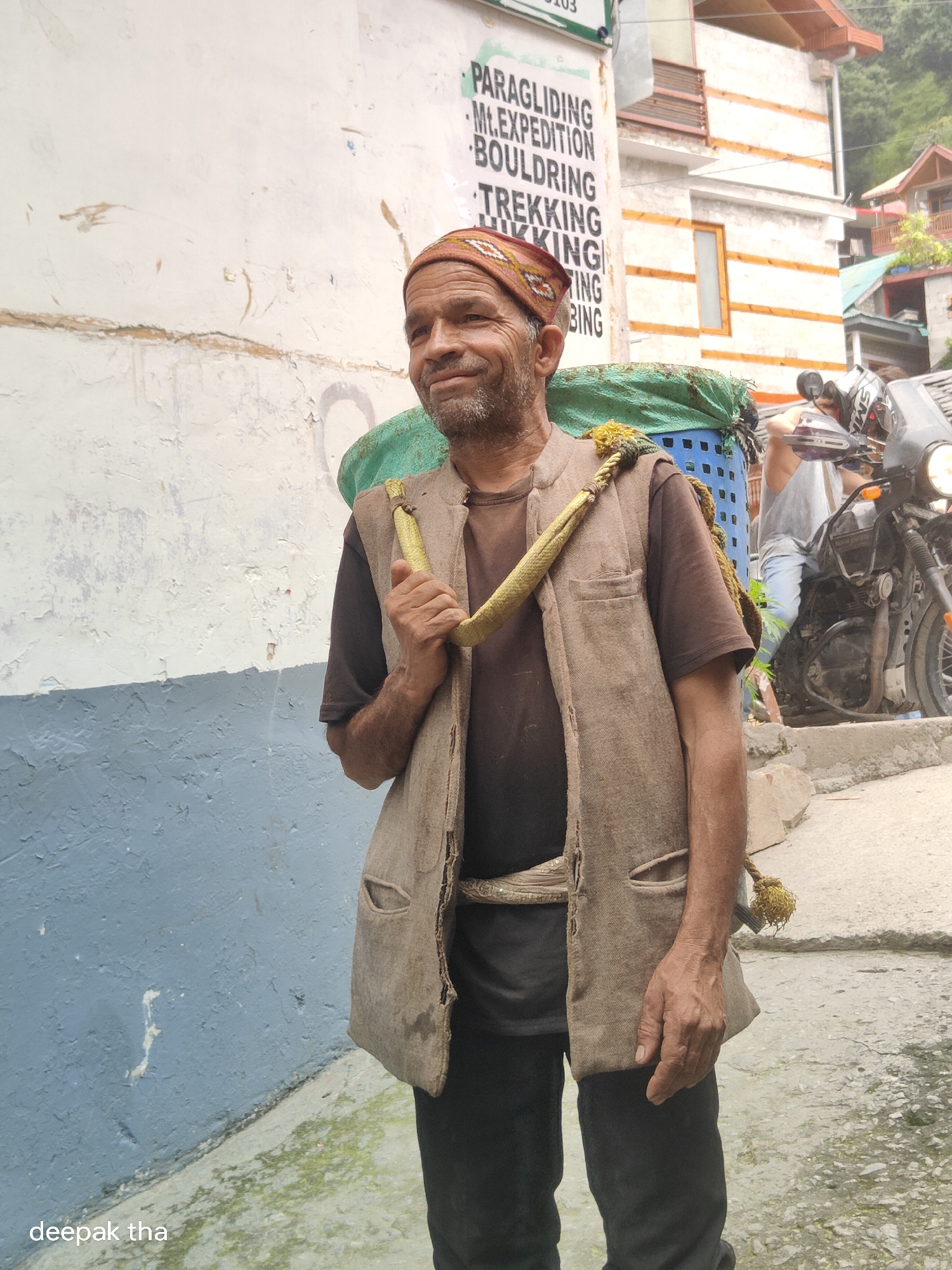 A man wearing a brown t-shirt, a tan vest, and a patterned head covering is carrying a large blue basket on his back. The basket is filled with green items and is secured with a rope. He is standing in an alleyway with a blue and white painted wall to his left. A motorcycle is parked on the right side of the frame. A sign on the wall lists various adventure activities such as paragliding, bouldering, and trekking. The image is labeled with "deepak tha" in the bottom left corner. The scene appears to be in Vashisht, India, a location known for its hot springs and trekking opportunities.