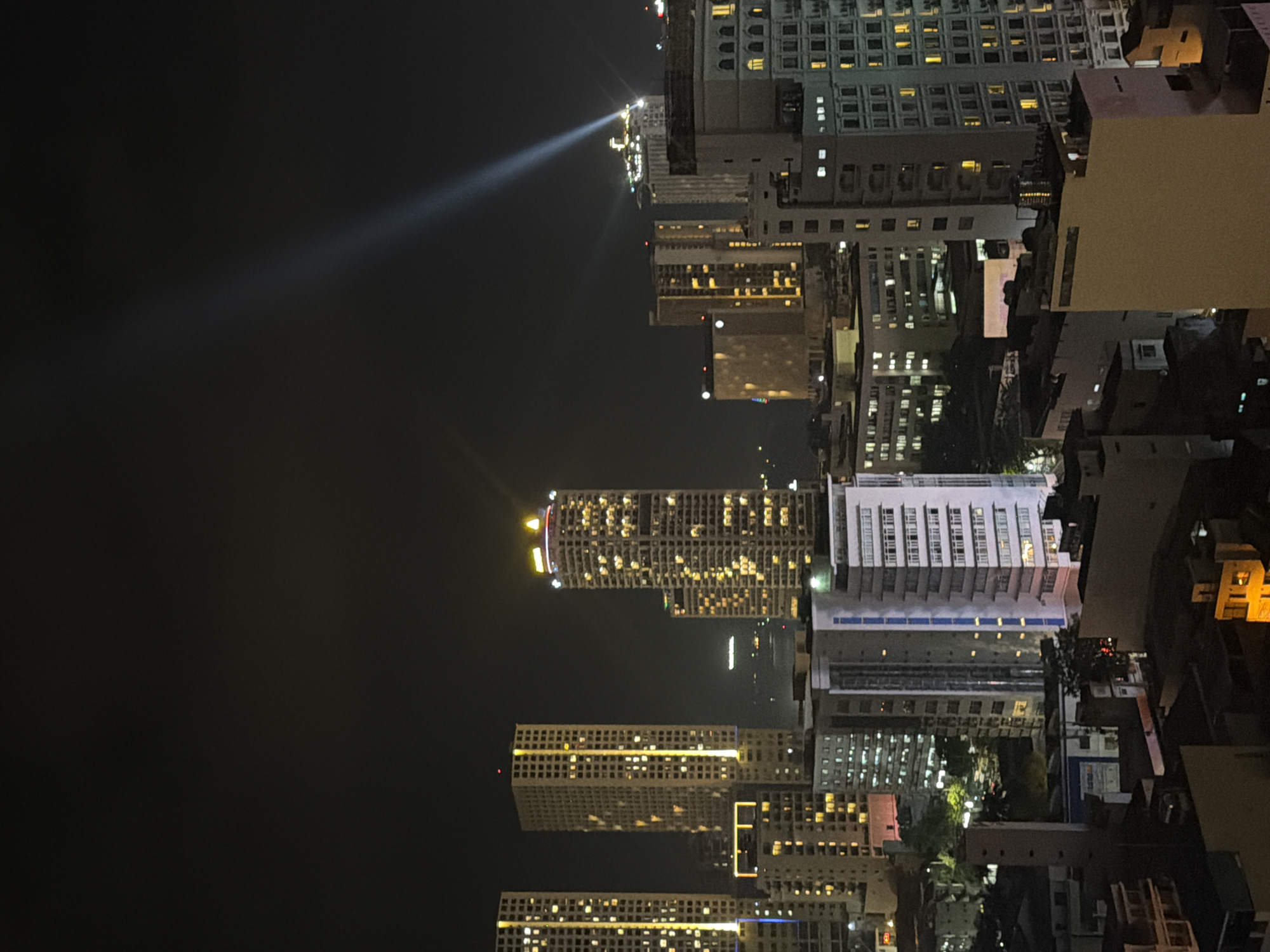 The image depicts a nighttime cityscape in Nha Trang Ward, Vietnam, characterized by numerous illuminated high-rise buildings under a dark, clear sky. In the midground, three prominent tall structures on the left feature numerous windows illuminated with yellow light. A slightly shorter building in front of the central tall structure also displays yellow illumination. To the right, another very tall building stands out with a distinctive illuminated yellow crown and multiple illuminated windows. Further to the right, a distant high-rise emits a strong, diagonal beam of white-blue light upwards into the sky from its rooftop. Scattered across the urban landscape are various other multi-story buildings, many with illuminated windows, contributing to the city's nighttime glow. Some structures display red lights on their rooftops, likely navigational or warning lights. A white and blue-striped building is visible in the lower-midground left. The immediate foreground reveals rooftops and upper portions of smaller structures with minimal illumination. No individuals or specific activities are discernible within the frame.