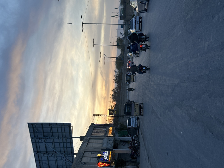 This image presents a street scene in Quetta City Tehsil, Pakistan, during either sunrise or sunset, evidenced by the multi-hued sky featuring orange, yellow, blue, and grey cloud formations.
The scene focuses on a wide, asphalt road displaying moderate wear. On the left, a two-story commercial building is visible. Its ground floor features an illuminated red and white sign for "Samwich," including a pizza slice graphic and the text "BELGIAN WAFFLES & PANINIS." Several motorcycles and a silver hatchback car are parked adjacent to the building. Above this structure, a large, unlit billboard framework is prominent against the sky.
On the roadway, traffic is present. In the mid-ground, a yellow three-wheeled auto-rickshaw, marked "GARDAN," moves away from the viewer. Behind it, three individuals on separate motorcycles are also traveling in the same direction. Further down the road, additional vehicles, including multiple cars and another auto-rickshaw, are observed. A row of streetlights with horizontal cross-arms, some adorned with small flags, lines the right side of the road. Beyond the road, an elevated earth embankment or hill with minimal vegetation is visible, leading to a distant horizon of hazy mountains.
