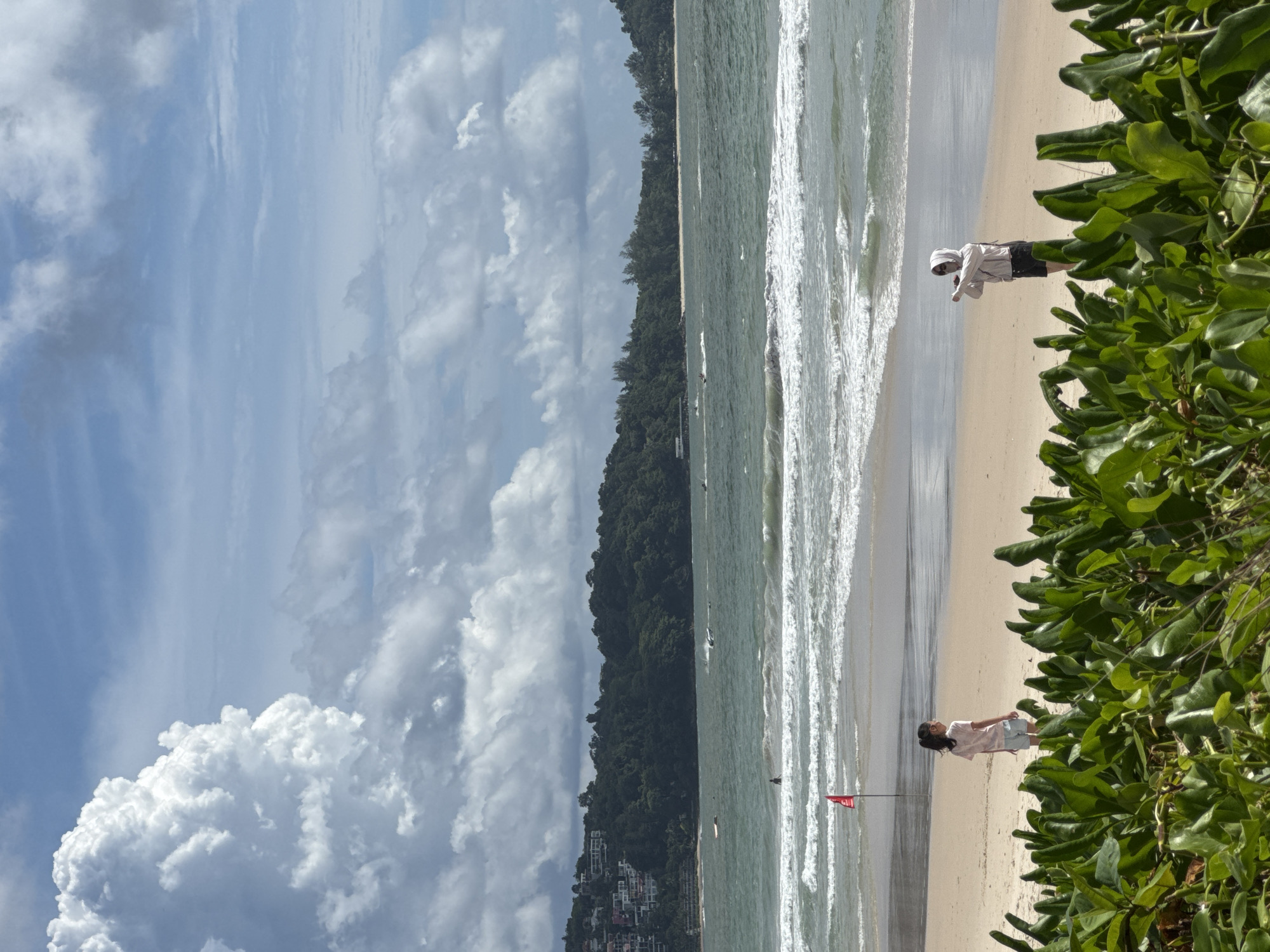 An eye-level, medium shot captures a serene beach scene on a cloudy day in Patong, Thailand. In the foreground, lush green foliage lines the bottom of the frame, with the sandy beach extending towards the ocean. A young girl in a light pink t-shirt and shorts stands on the left, facing the water. To her right, a person wearing a hooded jacket, sunglasses, and shorts is seen in profile, holding what appears to be a phone, possibly taking a photo. The ocean is a dynamic turquoise, with white waves crashing onto the shore. A red flag is planted in the sand near the waterline. In the middle ground, several jet skis are visible on the water. The background is dominated by a lush, tree-covered hillside dotted with buildings, characteristic of the Patong coastline. The sky is a dramatic mix of blue and white clouds, with a large, fluffy cloud formation prominent on the left.