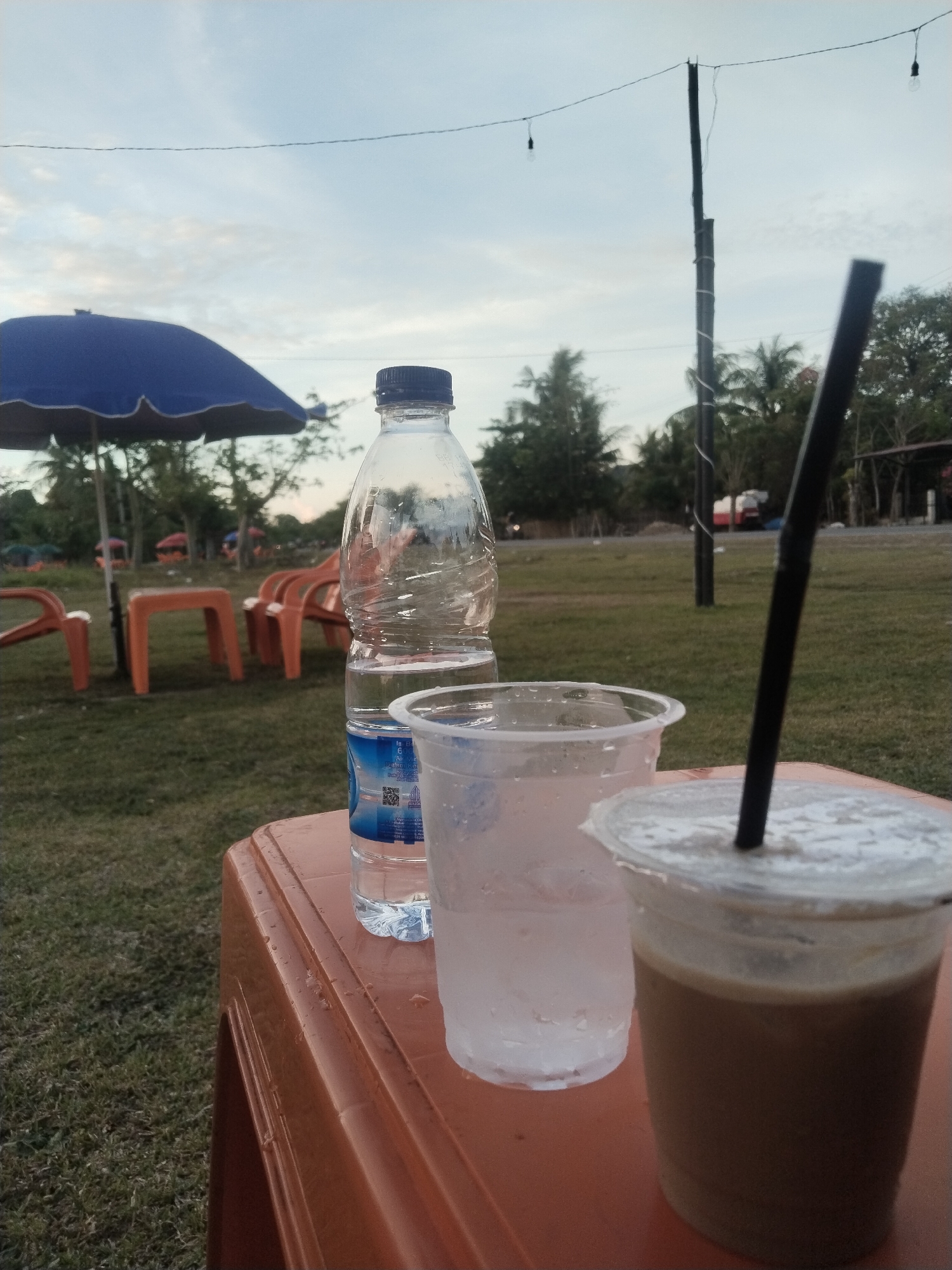 The image shows a close-up of a plastic table on a grassy surface, likely outdoors in Glokaron, Indonesia. On the table are three beverage containers: a partially empty bottle of water, a clear plastic cup with condensation, and a covered plastic cup with a straw, containing a coffee-colored drink. Behind the table are plastic chairs and tables with blue umbrellas, arranged on the grass. In the background, there are trees and utility poles, suggesting a park or recreational area. The sky is overcast, indicating it may be near dusk or dawn. There is no specific evidence of human activity visible besides the arranged furniture and drinks.