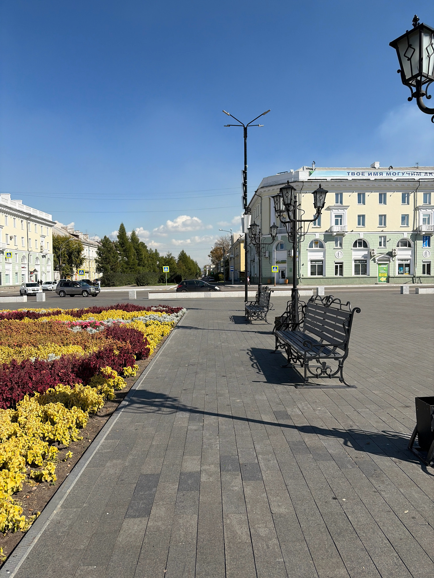 A sunny, clear day in Angarsk, Russia, is depicted in this photograph taken from a paved plaza. In the foreground, a flowerbed showcases a vibrant mix of red and yellow foliage. The plaza itself is paved with grey rectangular stones. Several ornate black park benches are visible, casting shadows on the pavement, along with streetlights and a trash receptacle. Across the plaza, a multi-story building with a light yellow facade and green accents stands under a bright blue sky. The building features numerous windows and some signage. In the background, trees and other buildings line a street where cars are visible, including a grey SUV and a white sedan. Pedestrian crossings are marked with yellow warning signs.