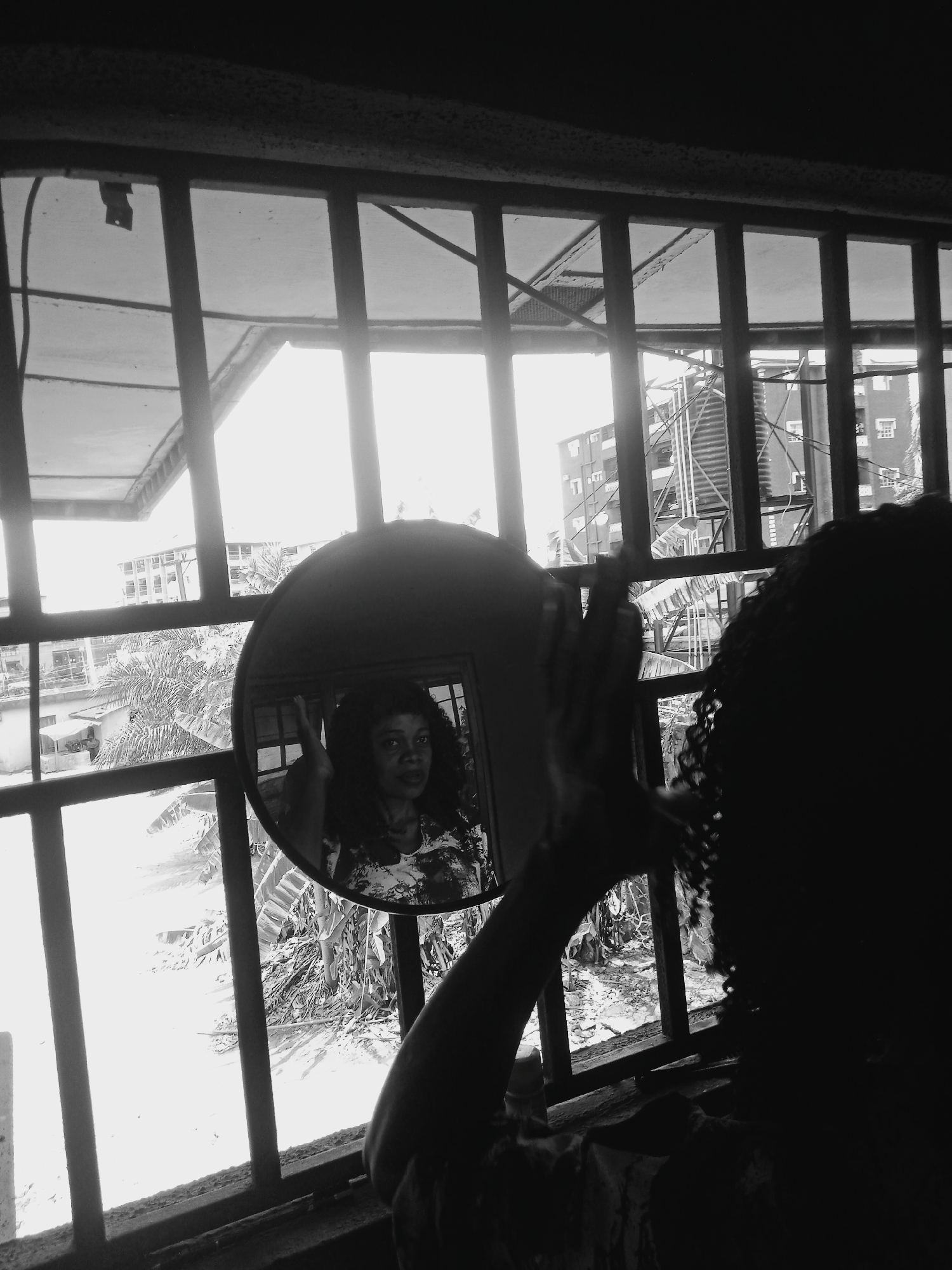 A black and white, medium shot depicts a woman standing in front of a barred window, holding a circular mirror that reflects her face. The woman facing away from the camera is in silhouette, with curly hair visible. The mirror shows a woman with darker skin tone, looking directly at the viewer, wearing a light-colored floral patterned top. Beyond the window bars, a cityscape is partially visible. Buildings, trees, and what appear to be water tanks are seen through the window. Based on the style of architecture and environment, it is assumed that the image was taken in the city of Onitsha, Nigeria.