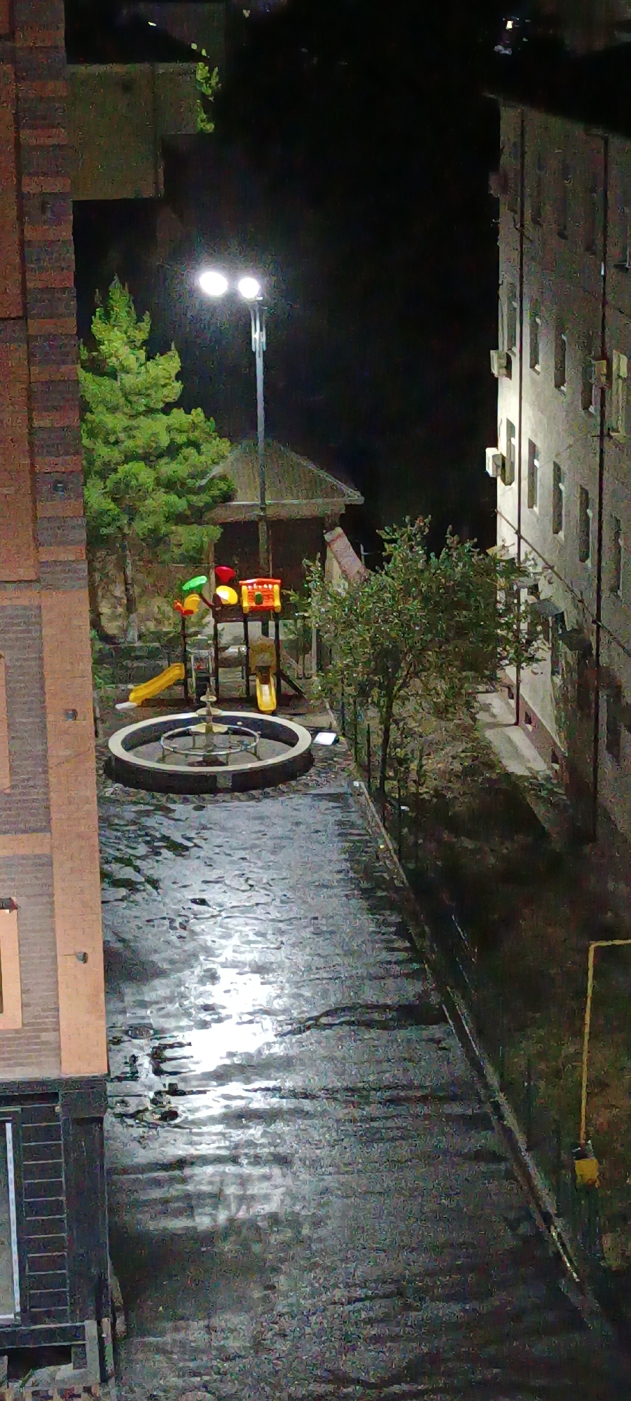 The image, taken at night in Fergana City, Uzbekistan, depicts a courtyard area between two apartment buildings. A playground structure with colorful slides and a red and yellow tower is visible, situated near a circular fountain. A street lamp illuminates the scene, casting light on a wet pavement. The ground appears to be wet, possibly from rain or recent watering, with reflections of the light and surrounding objects. One apartment building, constructed with brick on the left side of the image, is partially visible. The other apartment building, on the right, displays external air conditioning units and windows. Trees and shrubs are present in the courtyard, adding to the urban landscape. The overall atmosphere is dark and wet, typical of a city at night after precipitation.