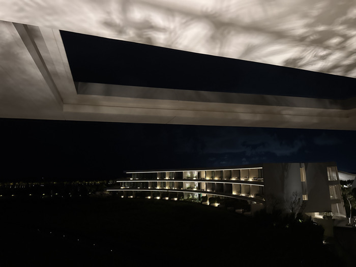 The image captures a modern building at night, seen from beneath a structure with intriguing light patterns. The building features illuminated balconies that curve gently. Below, a dark expanse suggests a lawn or field. Above, an overhang or roof detail shows shadows that resemble swirling clouds, creating a contrast between the geometric architecture and the soft, natural forms of the light play. The overall scene has a tranquil and mysterious ambiance.