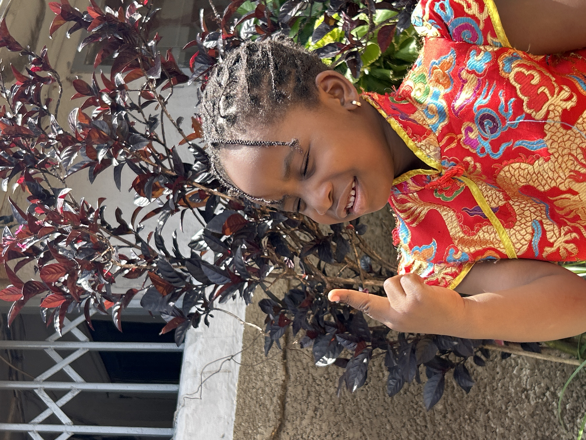 A young girl with braided hair and closed eyes smiles slightly as she holds up her index finger. She is wearing a red dress with gold dragon embroidery. The girl is standing outdoors in front of a brown wall with a metal gate and a plant with dark red leaves. The scene is located in Jos, Nigeria.