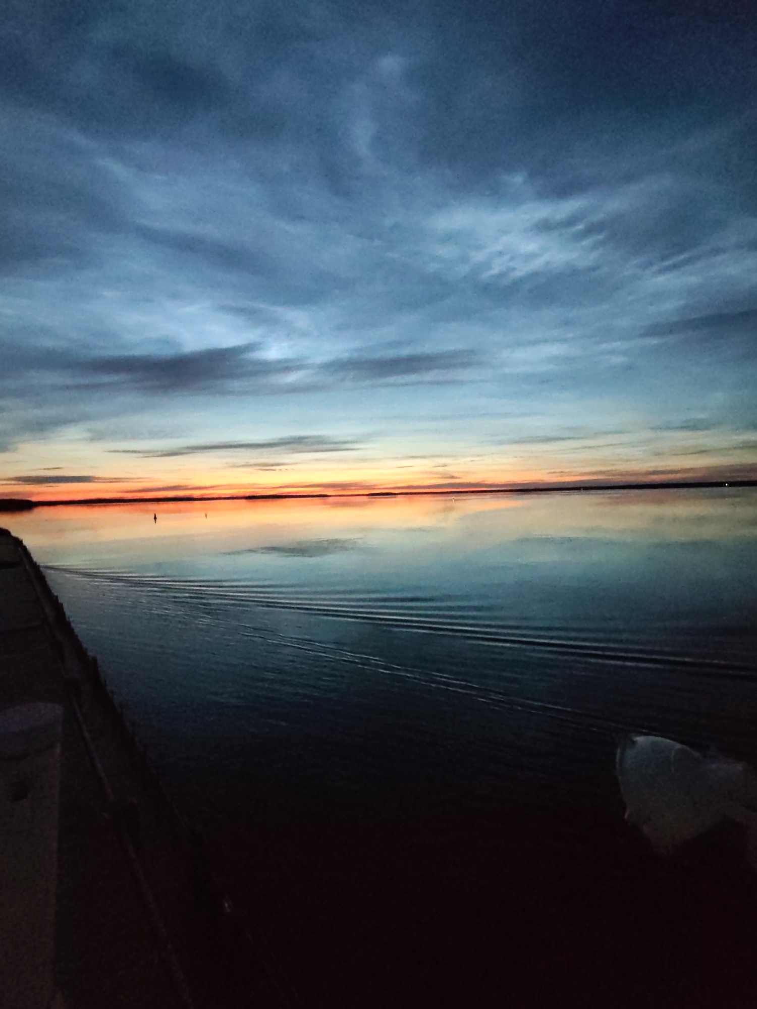 The image shows a calm body of water reflecting a twilight sky. The sky transitions from dark blue at the top to streaks of orange and pink near the horizon, indicating either sunrise or sunset. There are faint clouds scattered across the sky. The water is smooth, with a few subtle ripple patterns visible, suggesting a gentle disturbance.
Along the left side of the image, there is a dark, solid structure that appears to be a pier or embankment. Part of a white object, possibly a bollard or a light fixture, is visible on this structure. In the distance, two small, dark buoys are visible on the water.
The overall scene is serene and natural, with a focus on the interplay of light and water during the twilight hours. There are no visible people or specific landmarks that would definitively place the location within the city of Sheksna, Russia.