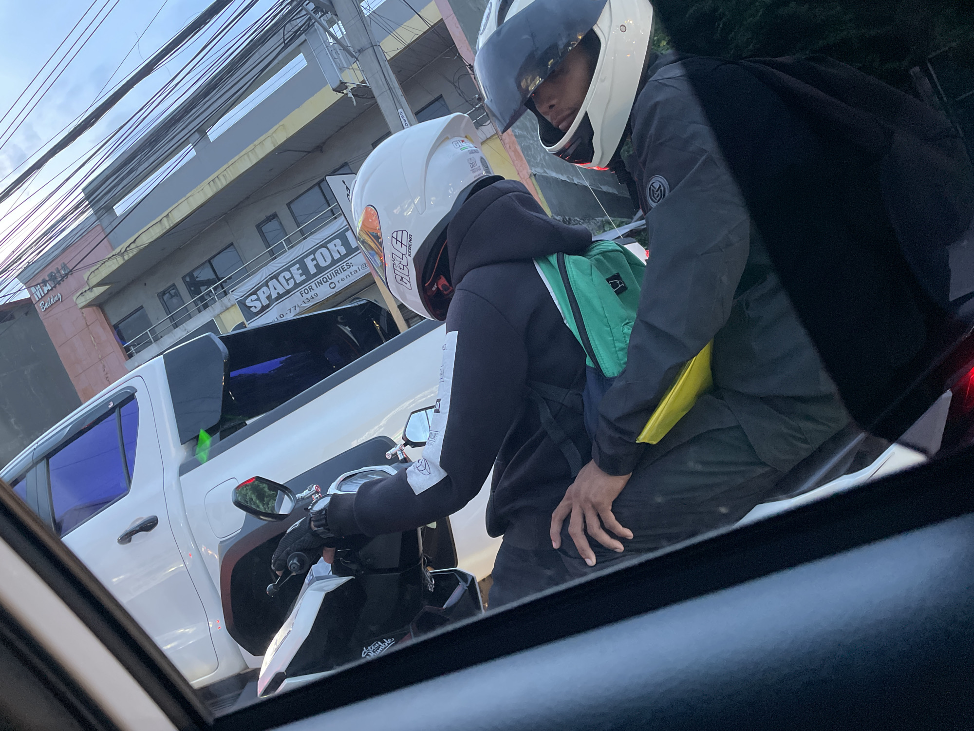 The image, likely taken from inside a vehicle, captures a scene in Davao City, Philippines. Two individuals on a white and black motorcycle are the central focus. Both are wearing helmets; the rider's helmet is white with orange accents, while the passenger's is white with a dark visor. The passenger is wearing a black jacket and carrying a green backpack with yellow accents. The rider is wearing a black hoodie. A white pickup truck with tinted windows is partially visible on the left. In the background, a building with a sign reading "SPACE FOR INQUIRIES" is seen, suggesting the location is an urban area with commercial properties. Power lines crisscross the sky.
