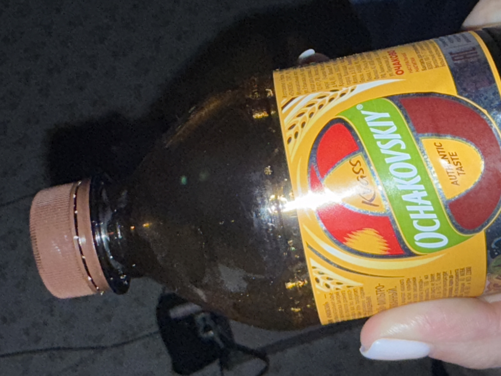 A close-up, canted shot depicts a plastic bottle of "Ochakovsky" kvas, a traditional Russian drink. The bottle is held by a person with light skin and white nail polish, the thumb visible near the label. The label is predominantly yellow, green, and red with wheat illustrations. The writing on the label is in Russian. The background is dark and blurry, suggesting an outdoor or low-light environment. The location of Mytishchi, Russia is not clearly evident in the picture.
