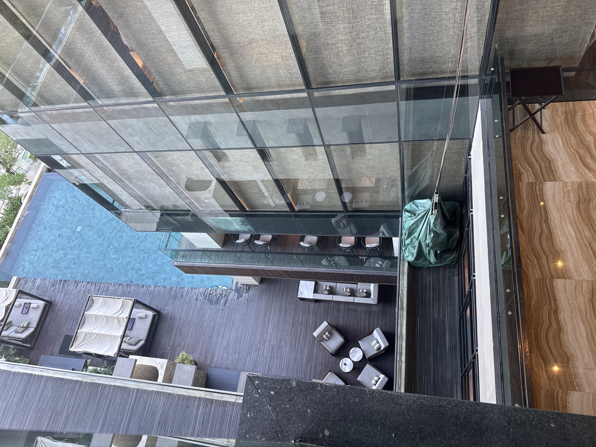 The image, taken from a high vantage point looking down, captures a luxurious outdoor area of what appears to be a modern hotel or resort. In the foreground, a dark, textured surface, possibly a balcony railing or floor, spans the bottom.
Below, a large, pristine blue swimming pool occupies the left side, reflecting natural light. Along the pool's edge, on a wooden deck, are several large, comfortable-looking sun loungers or daybeds, each featuring white fabric canopies and soft cushions. Some greenery in planters is also visible around this area.
Adjacent to the pool and extending towards the right is a spacious deck made of dark wood planks. This area is furnished with various outdoor seating arrangements. One section features a long, light-colored rectangular table surrounded by several chairs with light cushions, suggesting a dining or casual lounge space. Another section holds multiple armchairs with light grey cushions clustered around small, round white coffee tables, creating a more intimate seating arrangement. Further to the right, a large, dark green tarp covers an unknown object on the deck.
The backdrop consists of a modern building with a multi-story glass facade, reflecting parts of the sky and the surrounding environment. Behind some of the glass panels, a textured, light-colored fabric material, possibly sun blinds, is visible. On the far right edge, a section of a brown and beige marbled floor or wall texture is visible, along with a dark folding chair.
The scene is devoid of people, suggesting a tranquil and empty environment. The natural light indicates it is daytime. No visible text is present in the image. The setting suggests a high-end leisure destination, consistent with its given location context of Kuta Selatan, Indonesia.