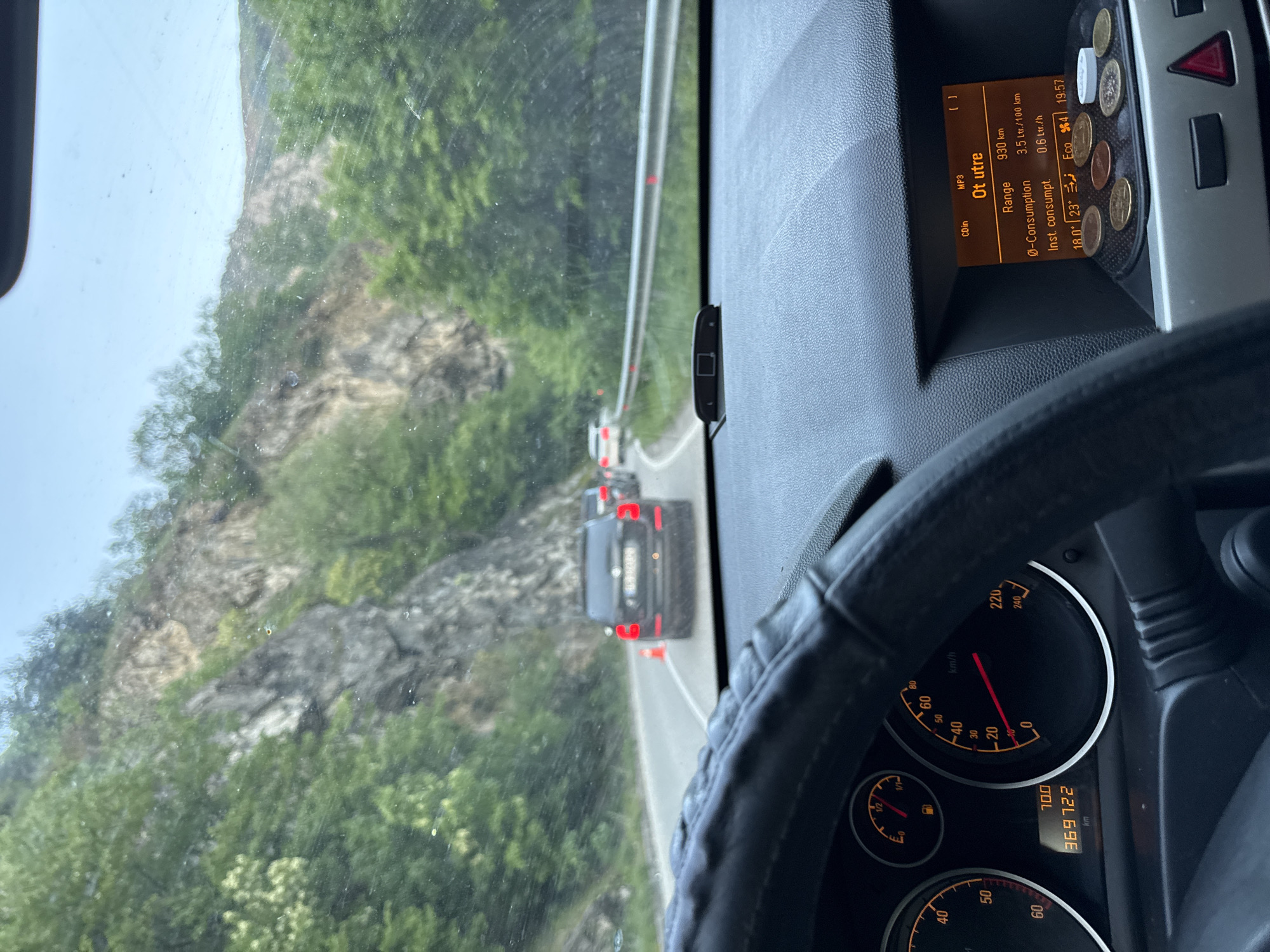 The image, taken from inside a moving vehicle, shows a winding road and mountainous landscape, likely in the vicinity of Бачковски манастир, Bulgaria, based on the dashboard display in Cyrillic script. The vehicle's dashboard is visible, displaying a range of 930 km, fuel consumption of 3.5 L/100km, and an instantaneous consumption of 0.6 L/h. The speedometer needle indicates a speed around 10 km/h, and the odometer reads 369722 km. Coins are placed on the dashboard. Through the windshield, one can see a vehicle ahead, marked by orange cones, suggesting road work or traffic control measures. The exterior landscape is dominated by a rocky hillside covered with green vegetation.