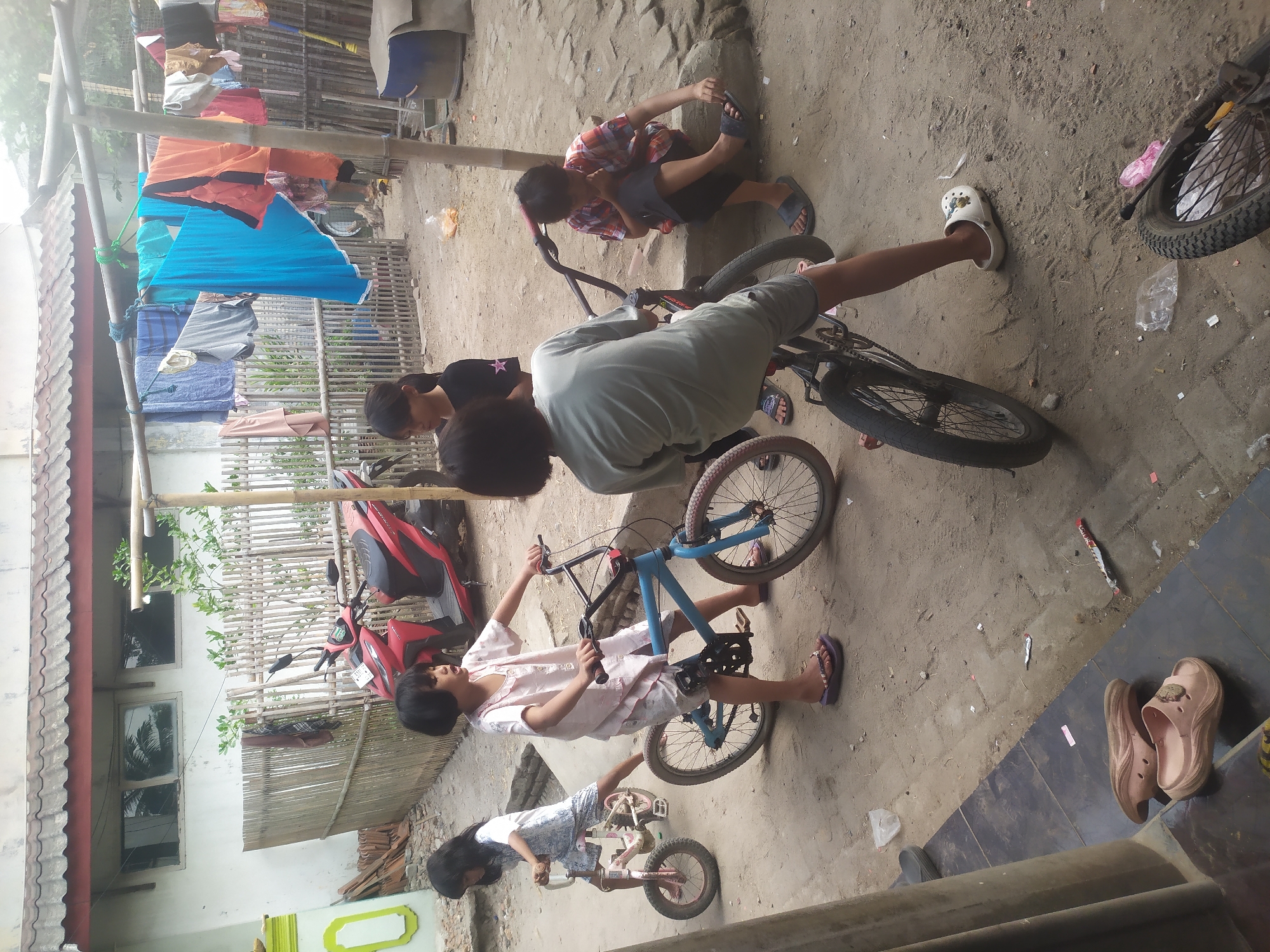 In Kragilan, Indonesia, a group of five children are gathered in a dusty, open yard with a building and bamboo fence in the background. Four children are interacting with bicycles. One girl in a white and blue patterned dress is on a pink bicycle, while another girl in a pink shirt and shorts is on a blue bicycle. Two other children are partially visible, one standing and looking towards the bicycles, and another sitting on a rock. A red motorcycle is parked behind a bamboo fence. Clothes are hanging on a clothesline above the scene. The ground is dirt and littered with debris.