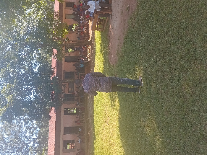 The image depicts an outdoor community gathering in Idakho South ward, Kenya. In the foreground, a male subject, viewed from the back, stands on a green grass lawn. He wears a dark, patterned long-sleeved shirt, dark blue jeans, and light-colored shoes. He faces a crowd of approximately 50-70 individuals, comprising men, women, and children, gathered in the midground. This crowd is seated on benches and directly on a reddish-brown dirt area in front of a single-story building. The building features light-colored walls, a reddish corrugated metal roof, and multiple open rectangular windows and doorways. Several large, leafy trees are positioned between the speaker and the building, partially obscuring parts of the assembled crowd and the structure. In the distant background, a dense stand of tall, slender trees is visible under a bright sky. The scene is illuminated by natural daylight, casting distinct shadows. The central figure appears to be addressing the assembled group.