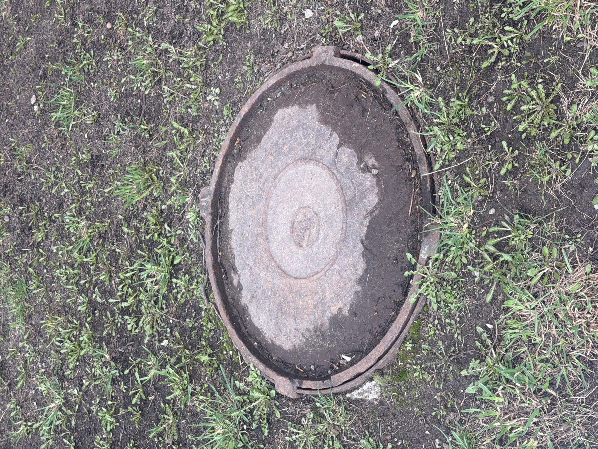 The image shows a circular, rusty metal manhole cover surrounded by soil and sparse green vegetation, likely grass and weeds. The manhole cover appears to be slightly recessed, with a dirt-filled depression on its surface, particularly around the concentric circular design in the center. The environment suggests an outdoor location, potentially a park or green space in a residential area. The general disrepair and weathered appearance of the manhole cover indicate some level of neglect or lack of recent maintenance. There is no evidence of human presence or activity in the frame. Location: Chaikovsky, Russia.