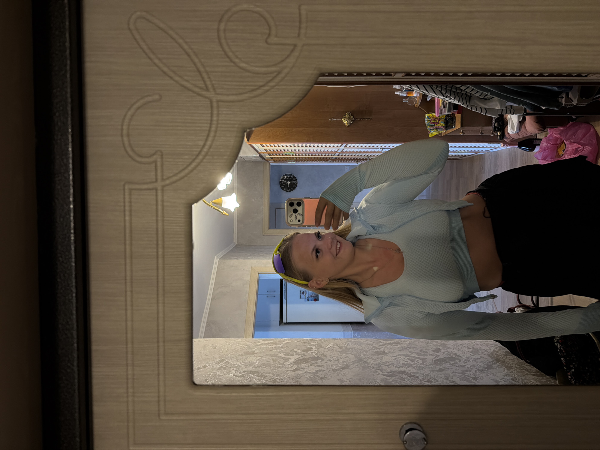 A young woman with blonde hair and a bright smile, wearing a light blue athletic outfit, poses for a mirror selfie. She is in what appears to be a bedroom or dressing area, with a mirror covering most of the frame. The mirror reflects a cluttered room with a wardrobe, a refrigerator, and a clock on the wall. The woman is holding a pink smartphone in her right hand, angled to capture her reflection. She is also wearing yellow-tinted sunglasses on her head. The room's decor is somewhat dated, with textured wallpaper and wooden furniture. The overall impression is of a candid moment captured at home.