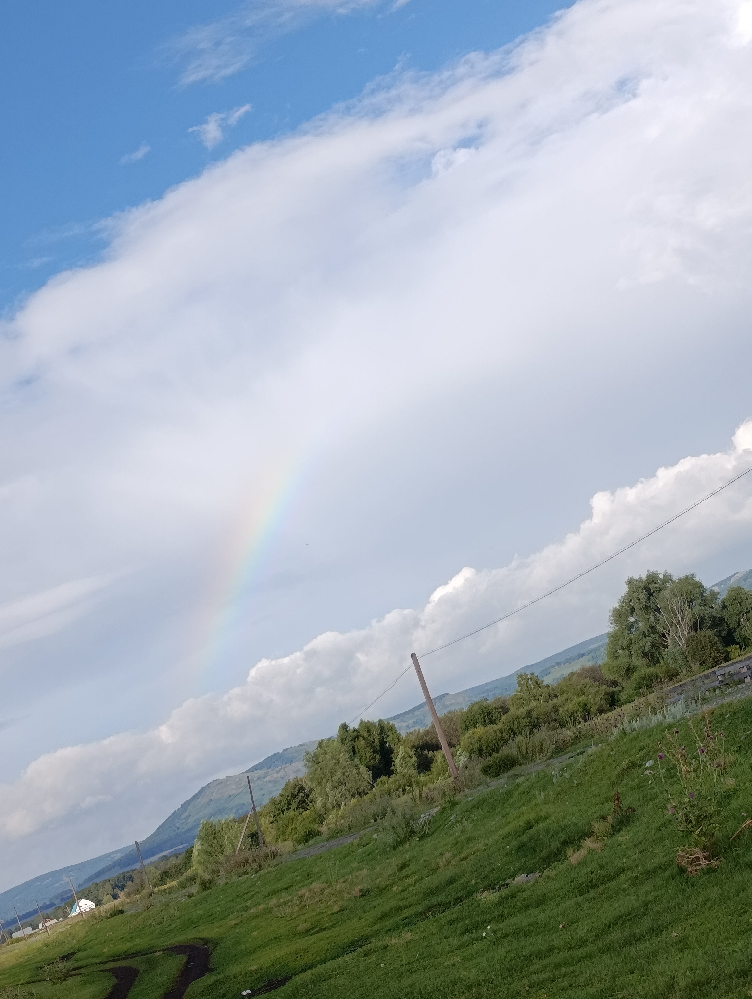 This is a landscape photograph, taken outdoors in a rural setting that could be the outskirts of Нижнетагирово, Russia. The image is dominated by a sky filled with dramatic clouds, with a faint rainbow visible on the left side. In the midground, a rolling green hill slopes upwards, covered in grass and sparse vegetation. Various trees and bushes are scattered across this hillside, with some appearing denser than others. A few utility poles, some with visible wires, are sparsely placed. In the background, a range of layered, bluish-green mountains stretches across the horizon. A small cluster of white buildings, possibly houses, is visible at the far left of the image, near the base of the hill. The overall lighting suggests it is daytime, with a mix of sunlight and cloud cover.