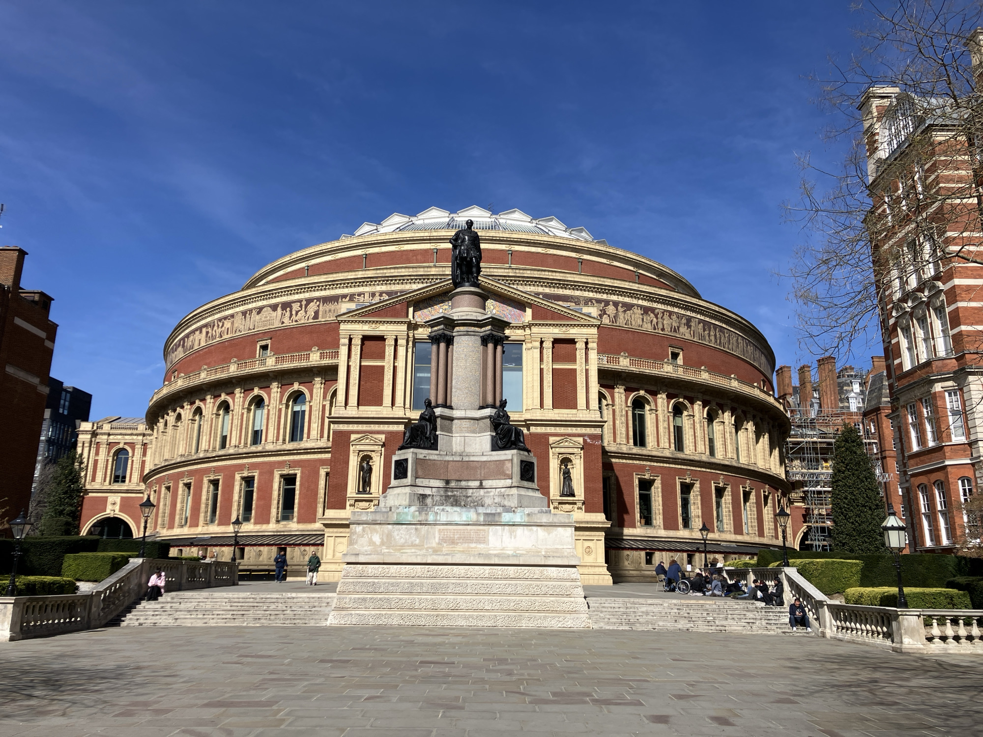 The image depicts the Royal Albert Hall, a large, circular, red-brick building with extensive yellow-cream terracotta detailing, situated in South Kensington, London, United Kingdom. The building features a prominent frieze depicting figures, numerous arched windows, and a segmented glass and metal dome roof.
Centered in the foreground, directly in front of the Royal Albert Hall, stands a multi-tiered stone monument topped by a dark-colored statue of a male figure (Prince Albert) in ceremonial attire. The pedestal of the monument includes smaller seated figures.
A wide paved plaza of light-colored flagstones occupies the immediate foreground, leading to a set of symmetrical stone steps with balustrades on either side. These steps ascend to a raised platform surrounding the monument. Manicured green hedges line the edges of the raised platforms. Two black traditional streetlights with white shades are positioned near the balustrades.
Several individuals are present: a person in a pink top is seated on the left stairs. On the right side, a group of people is seated on the stairs and raised platform, one individual in a wheelchair. Other people are standing or walking near the building's entrances.
To the right of the Royal Albert Hall, a multi-story red-brick building with white stone trim and multiple windows is partially visible, with scaffolding on a section further back. Bare tree branches are visible against the sky on the right. The sky is clear and bright blue with minimal cirrus clouds. The scene is illuminated by direct sunlight, casting shadows.